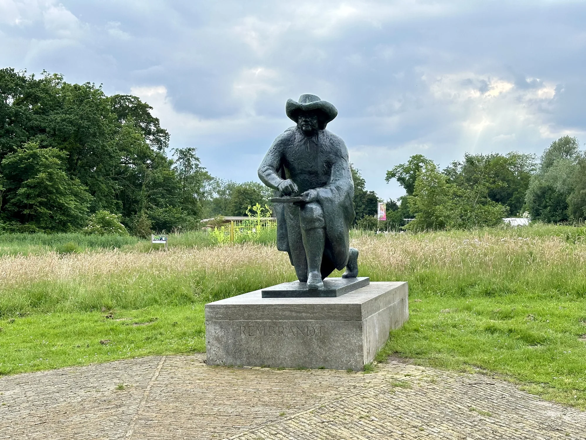 オランダのアムステルダムで撮影されたと思われる写真には、レンブラントの像が写っています。像は緑豊かな草地の中に立っており、背景には緑の木々が広がっています。空は曇り空で、少し陰鬱な雰囲気を醸し出しています。像は台座の上に立っており、台座には「REMBRANDT」と刻まれています。像自体は、帽子をかぶり、膝をついて何かを描いているような姿をしています。足元はレンガで舗装されています。全体的に、静かで落ち着いた雰囲気の写真です。