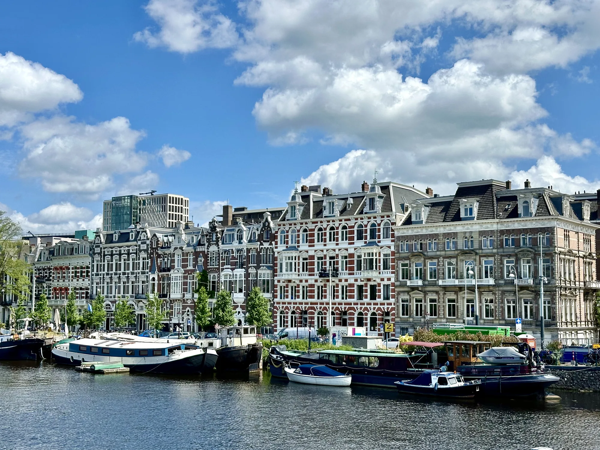 写真はオランダのアムステルダムで撮影された、運河沿いの風景です。穏やかな水面には、様々な種類のボートが停泊しています。運河沿いには、歴史を感じさせる美しい建物が立ち並び、その特徴的な外観が目を引きます。建物はレンガ造りで、白と赤茶色の装飾が施され、屋根の形もそれぞれ異なっています。建物の間には緑の木々が植えられており、景観に彩りを添えています。背景には近代的な高層ビルも見え、歴史的な景観と現代的な建築が調和している様子が伺えます。空は青く、白い雲が点在しており、穏やかで晴れた日であることがわかります。全体的に、アムステルダムの美しい街並みと運河の風景が調和した、魅力的な写真です。