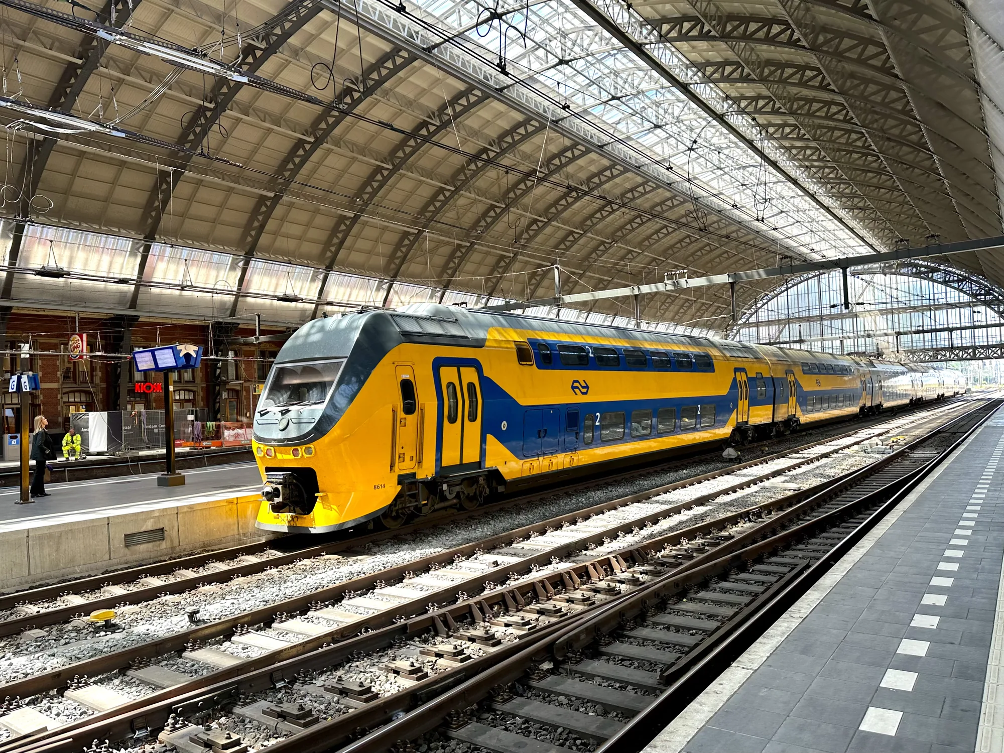 写真はオランダのアムステルダム中央駅で撮影されたものです。画面の中央には、黄色と青色のオランダ鉄道の二階建て列車が停車しています。列車の車体には大きな数字の「2」が描かれており、二等車であることを示しています。  駅のホームは灰色と白のタイル張りで、安全のために白い点線が引かれています。ホームの左側には、人影が二人見えます。一人は黒い服を着た女性で、もう一人は蛍光色の作業服を着た男性です。駅の建物はレンガ造りで、キオスクや店舗の看板が見えます。  駅の屋根はアーチ状で、金属製の骨組みとガラスが組み合わされており、自然光が差し込んでいます。レールは複数あり、複雑に分岐しています。全体的に、整然としていて明るい雰囲気の写真です。