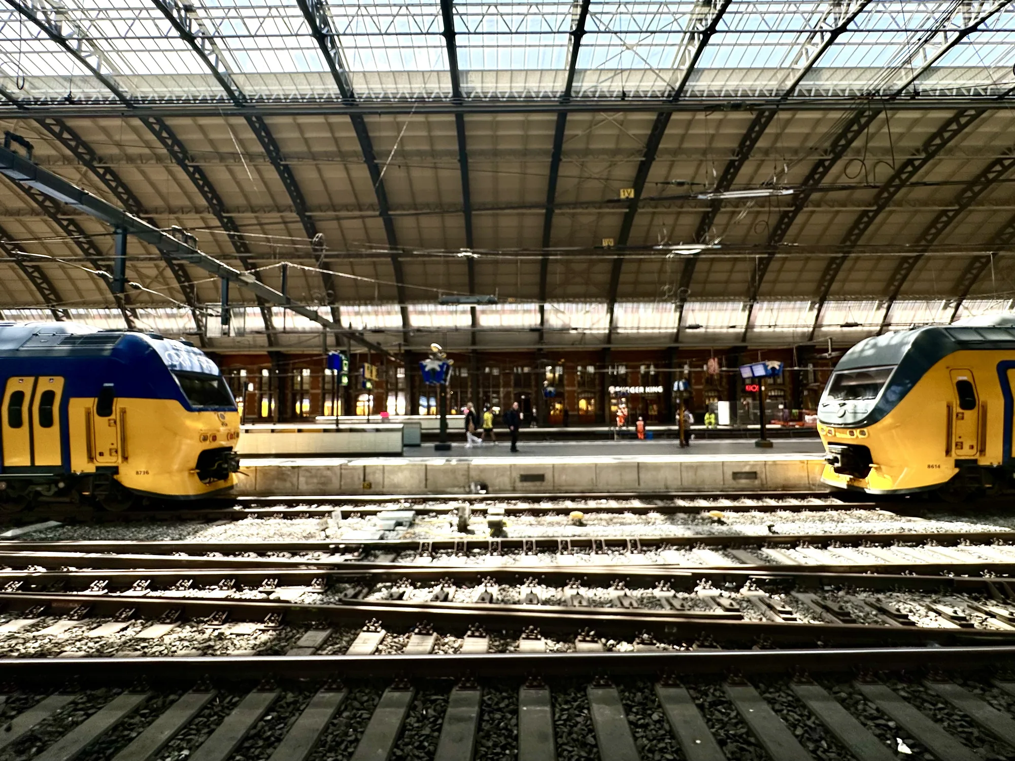 写真は、オランダのアムステルダム中央駅のプラットフォームを捉えています。画面の両端には、特徴的な黄色と青色のオランダ鉄道の車両が停車しています。プラットフォーム上には数人の人が歩いており、駅構内には店舗やレストランのような建物が見えます。駅の屋根は鉄骨構造で、一部がガラス張りになっており、自然光が差し込んでいます。線路には砂利が敷かれ、複数のレールが並んでいます。全体的に、活気がありながらも落ち着いた雰囲気の、ヨーロッパの駅の日常風景を写し出しています。