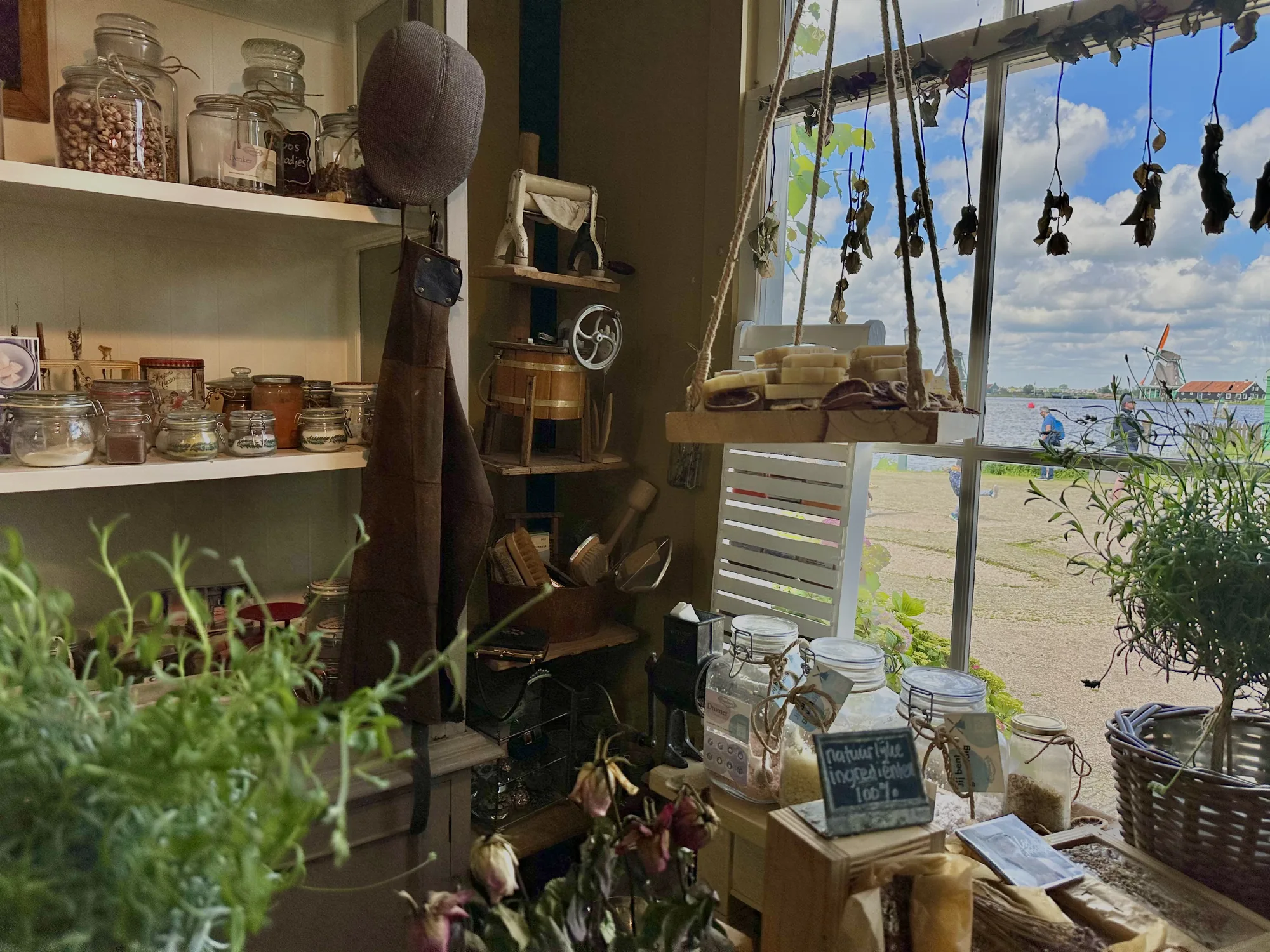 オランダのアムステルダムで撮影された写真には、可愛らしい雑貨店の店内の様子が写っています。画面左側の棚には、ナッツやスパイス、ジャムなどが瓶詰めされて整然と並べられています。棚の横には、帽子と革のエプロンが掛けられており、その下には昔ながらの洗濯機や、シャベルなどの道具が置かれています。窓際には、ロープで吊るされた棚があり、石鹸やドライフルーツなどがディスプレイされています。窓からは、運河と風車が見え、オランダらしい風景が広がっています。全体的に、温かみのある照明と、木製の家具や雑貨が、ノスタルジックで落ち着いた雰囲気を醸し出しています。前景には観葉植物があり、写真に奥行きを加えています。