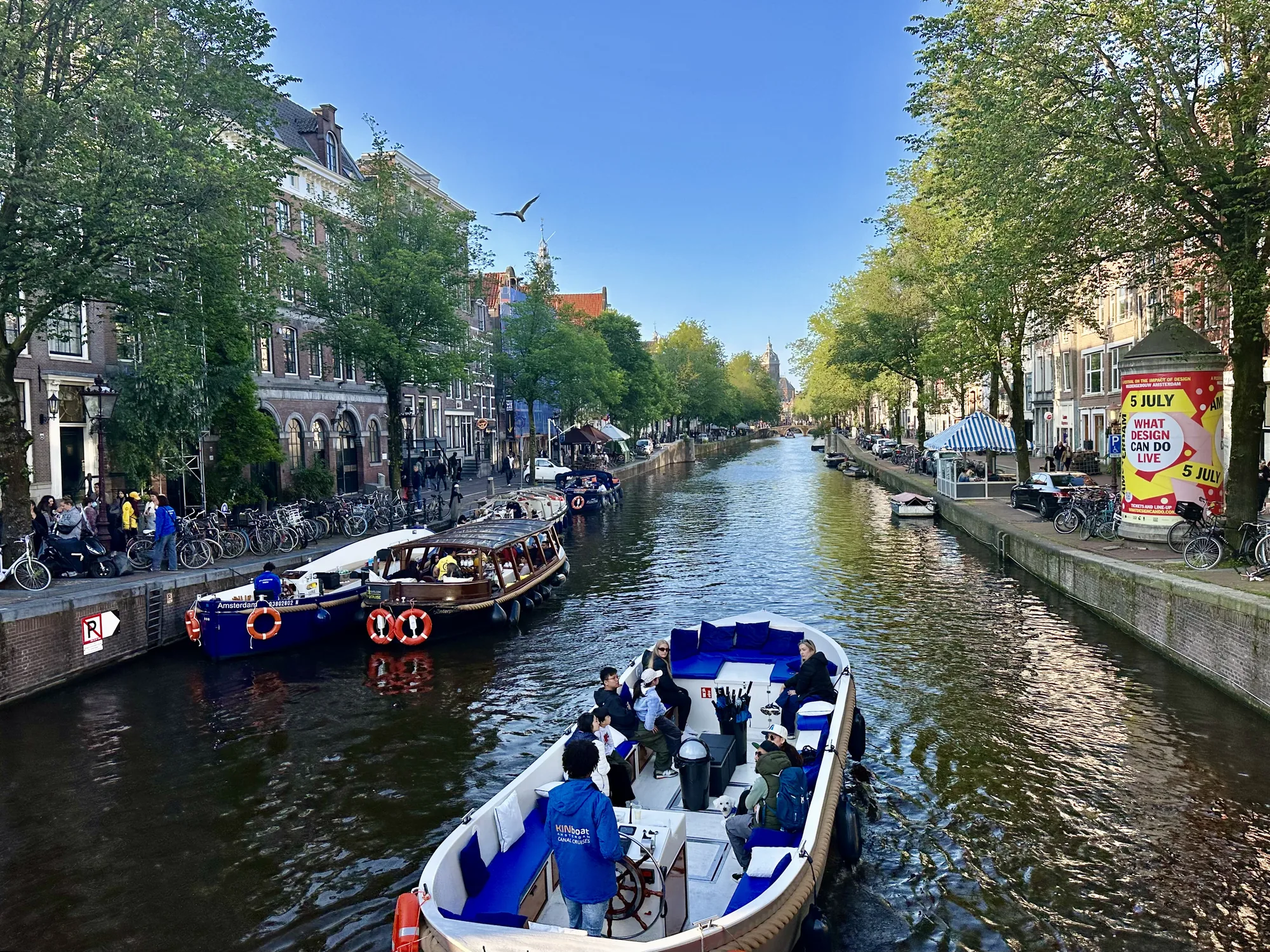 オランダのアムステルダムで撮影された写真です。運河を行く観光ボートからの視点で、運河の両側に立ち並ぶ建物と水面が写っています。運河には複数のボートが航行しており、手前のボートには複数の乗客が乗船しています。運河沿いには、レンガ造りの建物や木々が並び、人々が歩いたり、自転車に乗ったりしている様子が見られます。建物の壁には「5 JULY WHAT DESIGN CAN DO LIVE」と書かれたポスターが貼られています。空は晴れており、鳥が1羽飛んでいます。全体的に穏やかで観光地の活気を感じさせる雰囲気です。