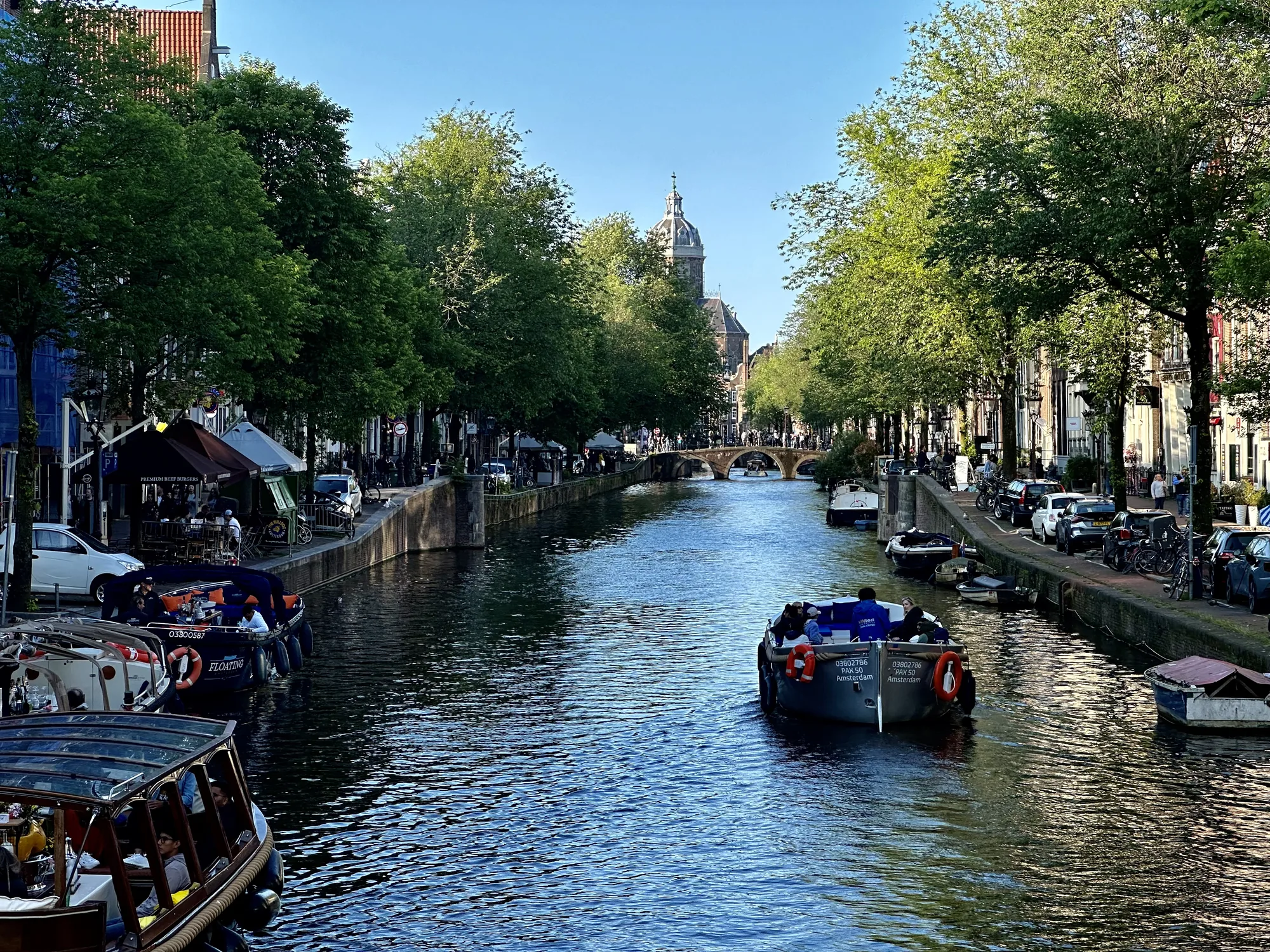 写真はオランダのアムステルダムで撮影された、運河沿いの風景を捉えたものです。青く穏やかな運河が画面の中央を貫き、その両側には緑豊かな街路樹が並んでいます。運河には観光客を乗せたボートがいくつか浮かび、賑やかな雰囲気を醸し出しています。左側の岸辺にはカフェやレストランが軒を連ね、テラス席で食事を楽しむ人々の姿が見られます。右側の岸辺には車や自転車が駐輪され、人々の生活の息吹を感じさせます。遠景には橋が架かり、その先には教会の尖塔が見えます。空は晴れ渡り、全体的に明るく爽やかな印象を与える写真です。アムステルダムらしい、運河と街並みが調和した美しい景観が表現されています。