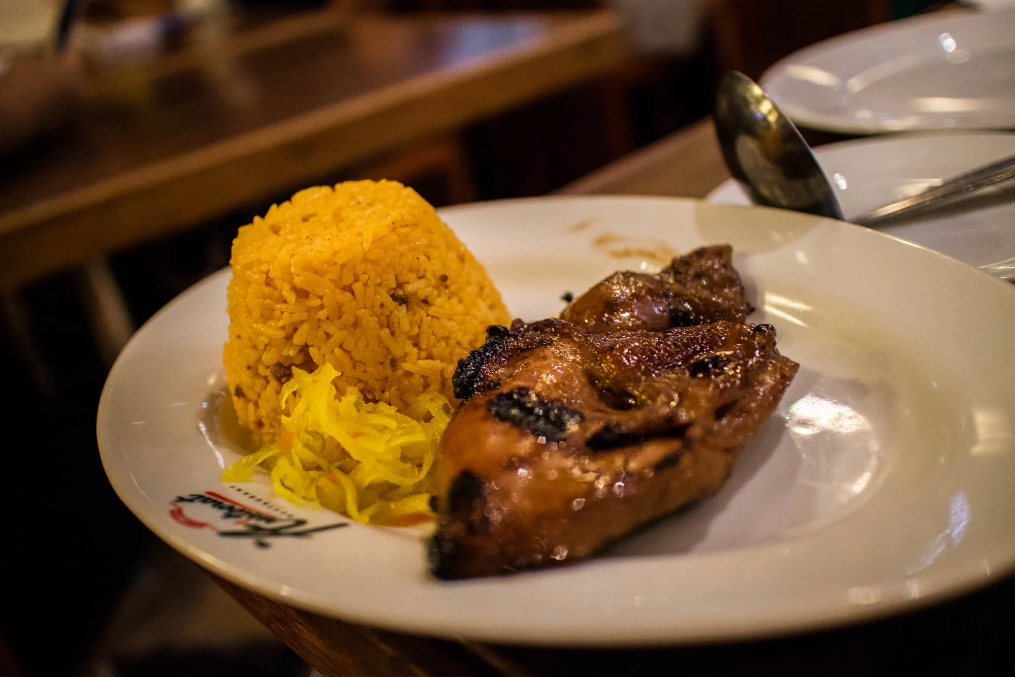 写真には、フィリピンのセブで撮影されたと思われる料理が写っています。白い皿に盛り付けられた料理は、こんもりと盛られた黄色いご飯、細切りの黄色い漬物のようなもの、そして照り焼き風の鶏肉です。鶏肉は焦げ目がついており、食欲をそそります。皿にはレストランのロゴらしきものがプリントされています。背景には木製のテーブルや積み重ねられた白い皿が見え、レストランの店内であることが伺えます。全体的に温かみのある照明で、美味しそうな雰囲気が伝わってきます。