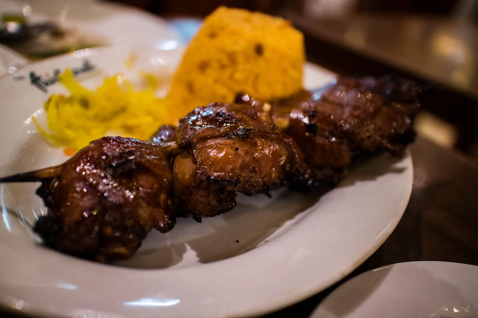 写真はフィリピンのセブで撮影されたと思われるBBQ料理のクローズアップです。メインは串に刺さった照り焼き風の肉で、濃い茶色のタレが塗られており、焼けた焦げ目が食欲をそそります。肉はジューシーで光沢があり、甘辛い香りが漂ってくるようです。  肉の串焼きの奥には、こんもりと盛られたオレンジ色のライスと、細切りの黄色い付け合わせが添えられています。ライスはターメリックライスか、アチョーテオイルで炊いたものかもしれません。黄色い付け合わせはパパイヤのピクルス（アチャラ）でしょうか。  料理は白い皿に盛り付けられており、皿の縁にはレストランのロゴらしきものが部分的に見えます。背景はぼやけており、テーブルの一部が写っています。照明は少し暗めで、料理にスポットライトが当たっているような雰囲気です。全体的に温かみのある色調で、食欲を刺激するような写真です。