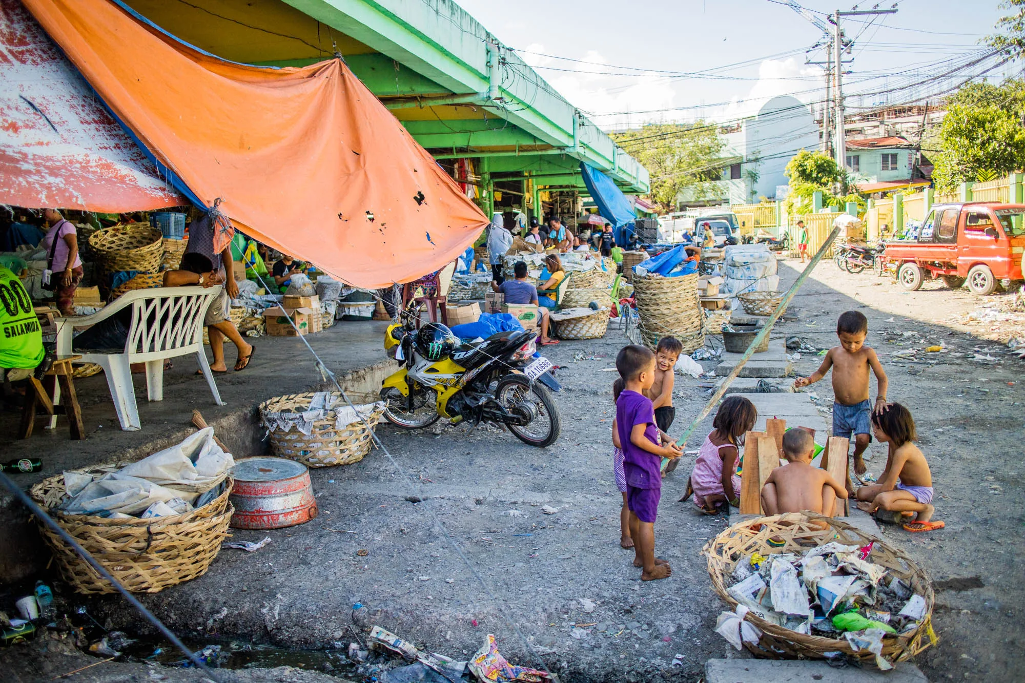 この写真はフィリピンのセブで撮影された、市場の一場面を捉えたものです。手前には裸足の子供たちが数人集まって遊んでいます。彼らの周囲には、ゴミが詰め込まれた大きな籠や、様々なものが散乱した路面が見られます。子供たちの後方には、黄色いオートバイが停められています。  さらに奥には、オレンジ色や青色のタープが張られた市場の屋台が並んでいます。人々が屋台の下で座っていたり、買い物をしていたりする様子が伺えます。市場には、商品を陳列するための大きな籠がたくさん積まれています。背景には建物や電線が写っており、生活感のある風景が広がっています。全体的に、市場の賑やかさと、子供たちの無邪気さが混ざり合った写真です。