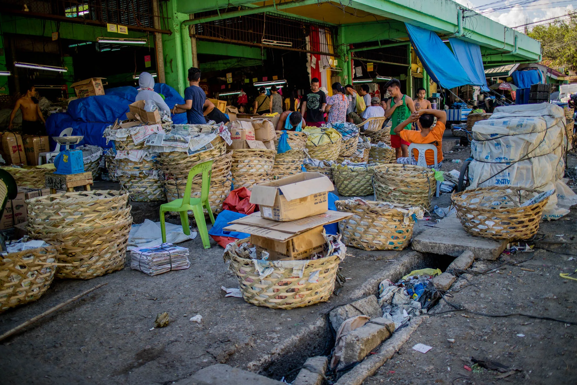 写真はフィリピンのセブで撮影された市場の様子を捉えています。手前には多数の竹製の籠が積み上げられており、中には新聞紙や段ボール箱などが入っています。奥には人々が行き交う市場の建物が見え、活気のある雰囲気が伝わってきます。床はコンクリートで舗装されており、排水溝にはゴミが溜まっています。全体的に、ローカルな市場の生活感あふれる光景が描写されています。