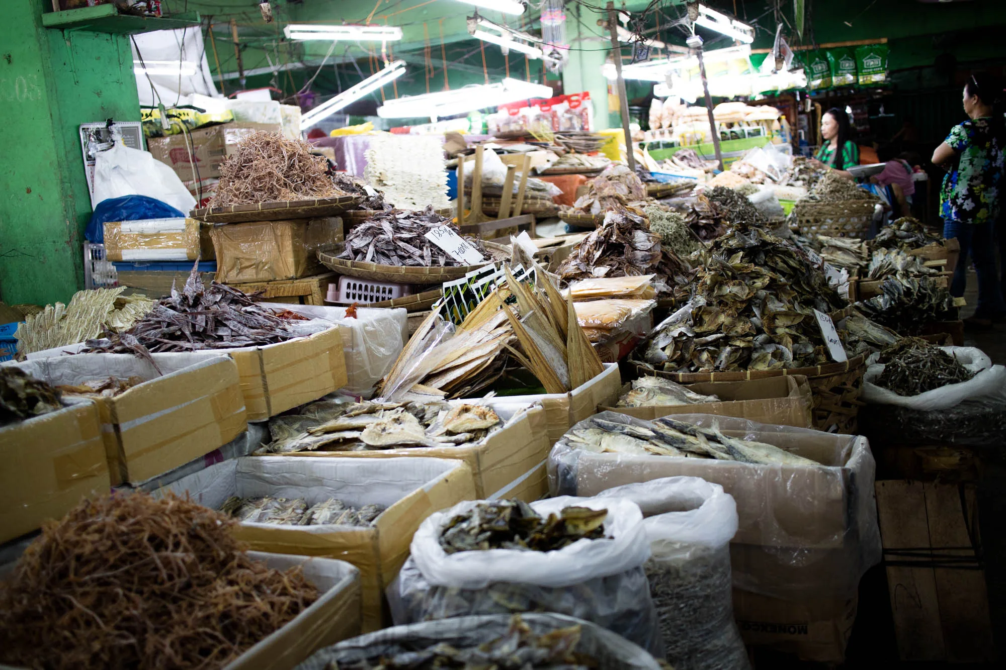 写真はフィリピンのセブ島で撮影された、賑やかな市場の一角を捉えています。特に目立つのは、大量の干物です。段ボール箱や竹籠に山積みされた様々な種類の干物が、所狭しと並べられています。干物の種類は様々で、細いものから平たいもの、丸ごと乾燥させた魚まで、多様な形状が見て取れます。色も、白っぽいものから濃い茶色まで、乾燥の度合いや魚の種類によって異なっているようです。  市場の雰囲気は活気に満ち溢れています。蛍光灯の光が全体を照らし、手前には干物を求める買い物客の姿も見えます。背景には、さらに多くの商品が並んでいる様子が伺え、市場の奥行きを感じさせます。雑多な商品が積み上げられている様子から、地元の人々の生活に密着した市場の様子が伝わってきます。