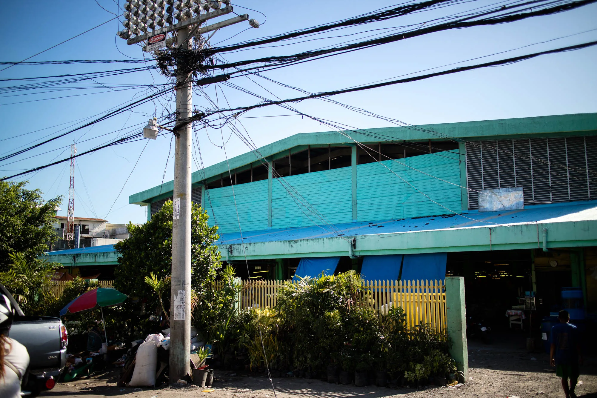 写真はフィリピンのセブで撮影されたもので、市場の周辺の風景を捉えています。空は晴れ渡り、電線が複雑に張り巡らされた電柱が印象的です。電柱にはメーターボックスや注意書きの看板が見られます。背景にはターコイズブルーの壁面を持つ建物が写っており、窓のような開口部にはルーバーが設置されています。建物の下には黄色い柵があり、植物が植えられています。道路脇には、商品が並べられた露店のようなスペースがあり、傘が立てられています。路面は舗装されておらず、人が歩いている様子も見られます。全体的に、セブの日常的な風景が写し出されており、ローカルな雰囲気を感じさせます。