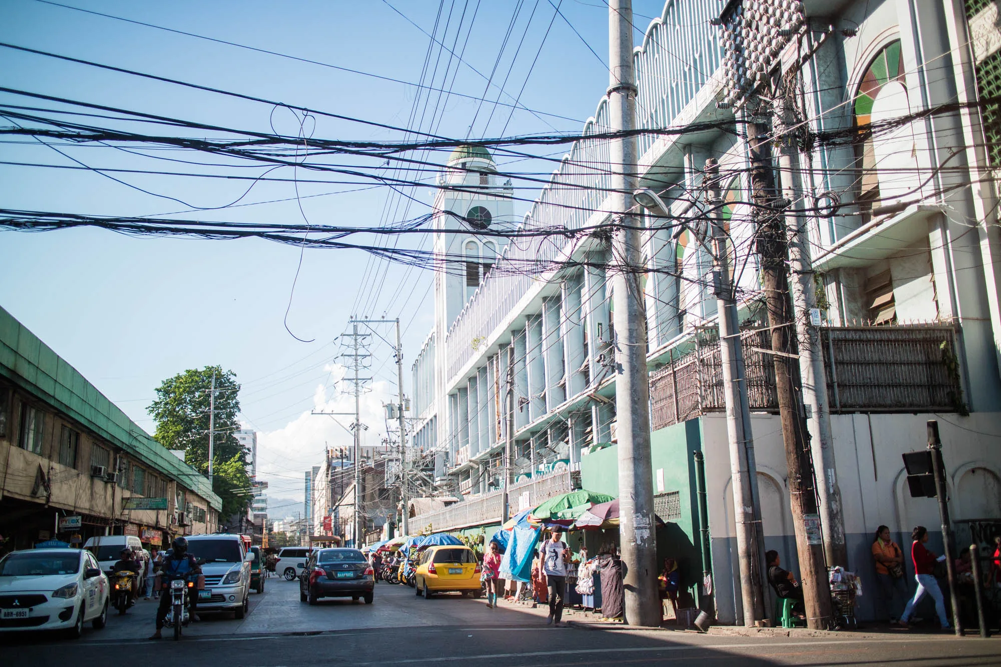 写真はフィリピンのセブで撮影されたもので、賑やかな街並みが写っています。手前には道路があり、車やバイクが走行しています。道路沿いには露店が立ち並び、人々が買い物を楽しんでいる様子が見られます。露店のカラフルな傘や商品が、活気ある雰囲気を醸し出しています。  背景には、特徴的な白い建物がそびえ立っています。建物の壁面には多数の縦長の窓が並んでおり、頂上には緑色のドーム型の屋根を持つ時計台が見えます。また、建物と電柱の間には、複雑に絡み合った電線が張り巡らされています。これは、東南アジアの街並みによく見られる光景です。  写真全体としては、セブの日常の風景を捉えたもので、生活感あふれる雰囲気を感じさせます。青空が広がり、日差しも強く、熱帯の気候が感じられます。