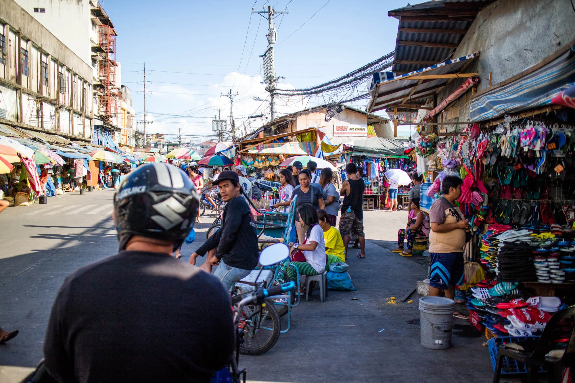 写真はフィリピンのセブ島で撮影された、活気のある市場の様子を捉えています。手前にバイクに乗った人物の後ろ姿があり、その先に多くの人々が行き交う様子が見えます。道路の両側には露店が並び、カラフルな傘や商品が所狭しと並べられています。電線が複雑に張り巡らされ、アジア特有の混沌とした雰囲気を醸し出しています。左側には古い建物が立ち並び、右側には簡易的な屋根が連なっています。人々は様々な服装をしており、中には商品を売る人々や、買い物を楽しむ人々がいます。全体的に日差しが強く、活気と熱気が伝わってくるような一枚です。