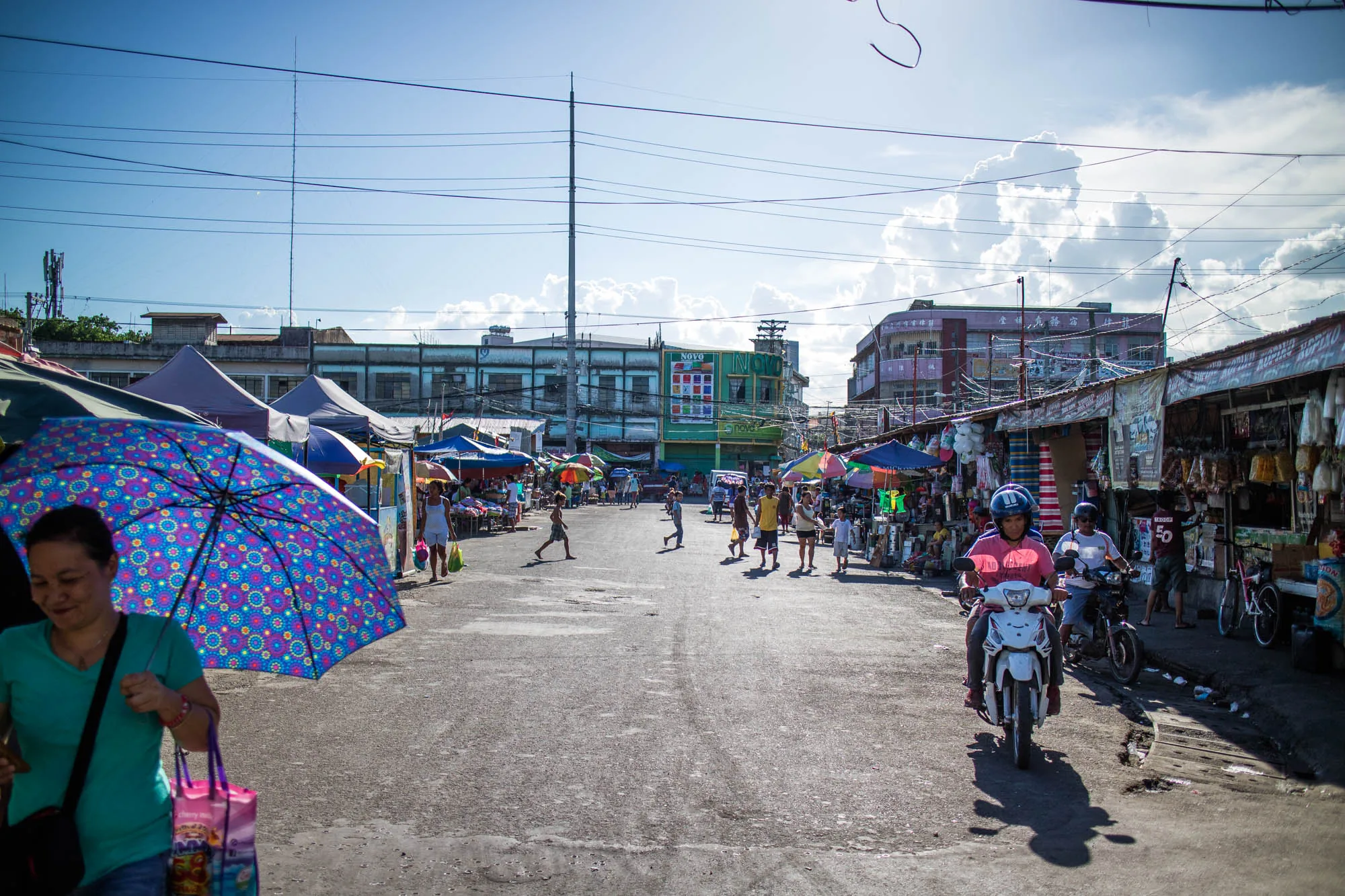 写真はフィリピンのセブで撮影された市場の様子を捉えています。中央には広い舗装された道があり、人々が歩いたり、子どもたちが走ったりしています。道の両側には屋台が並び、カラフルなパラソルやテントが日差しを遮っています。画面右側にはバイクに乗った人々が写っており、道の脇に停まっているバイクも見えます。建物は少し古びた様子で、電線が空中に張り巡らされています。空は晴れており、白い雲が浮かんでいます。活気があり、生活感あふれる市場の雰囲気が伝わってきます。