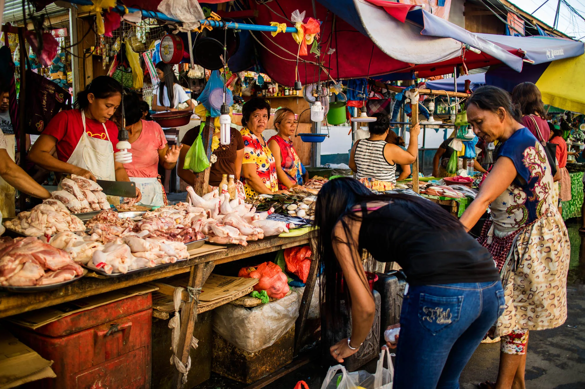 フィリピンのセブで撮影された写真には、活気に満ちた市場の様子が捉えられています。露店には、鶏肉や魚などの生鮮食品が豊富に並べられています。女性たちが店番をしており、肉を切り分けたり、客の対応をしたりしています。客は商品を吟味しており、買い物袋を手に取っています。市場の屋根には様々な色のタープや装飾が施されており、活気と賑わいを演出しています。電球が吊り下げられ、夕暮れ時の市場を照らしています。写真全体から、東南アジアの市場の喧騒と人々の生活感が伝わってきます。