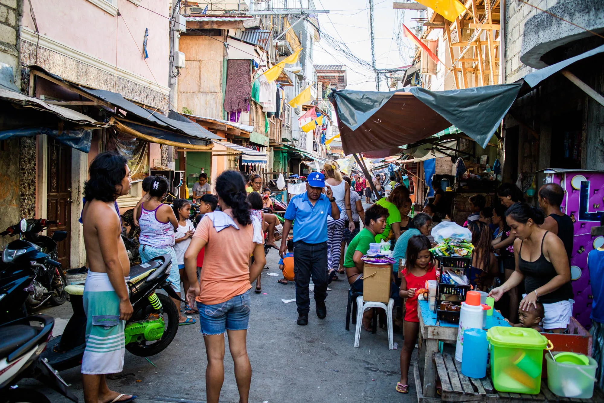 この写真はフィリピンのセブで撮影された、賑やかな街並みの様子を捉えています。画面中央には、青いポロシャツを着た男性がこちらに向かって歩いており、周囲には多くの人々が様々な活動をしています。  背景には、密集した建物が立ち並び、屋根や壁にはトタン板や木材が使用されているのが見えます。建物と建物の間には、洗濯物や旗のようなものが吊るされ、生活感があふれています。電線が複雑に張り巡らされ、ごちゃごちゃとした雰囲気を醸し出しています。  画面左側には、オートバイが数台停められており、その手前には上半身裸の男性が立っています。また、子供たちが走り回っていたり、露店のような場所で人々が食事をしていたりする様子が写っています。露店には、飲料や食品が並べられており、女性がそれらを提供しているようです。全体的に活気があり、人々の生活の息吹が感じられる写真です。