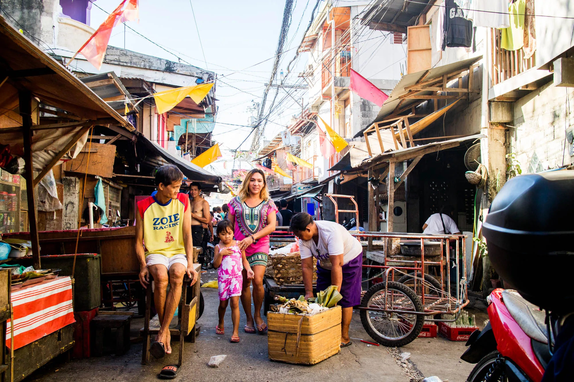 写真はフィリピンのセブで撮影されたもので、活気のあるストリートの様子を捉えています。中央には、ピンクのドレスを着た母親とその隣にピンクの花柄のワンピースを着た幼い女の子が写っており、手をつないで歩いています。母親は笑顔で、娘は少し緊張した面持ちです。写真左手には、黄色と赤のシャツを着た男性が椅子に腰掛けており、リラックスした様子です。彼の背後には、屋台のようなものがあり、様々な商品が並んでいます。右手には、白いシャツを着た男性が木箱を覗き込んでおり、その横には三輪車が置かれています。  背景には、電線が複雑に張り巡らされており、カラフルな旗が飾られています。建物の多くは簡素な造りで、一部は老朽化しているようにも見えます。洗濯物が干されていたり、剥き出しの木材が使用されていたりと、生活感があふれています。路上には、ゴミや瓦礫が散見され、街の喧騒が感じられます。光は強く、日中の暑さを伝えています。全体的に、セブの庶民的な生活を垣間見ることができる写真です。