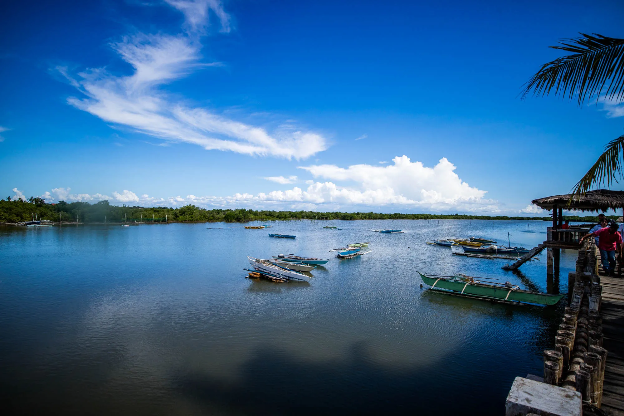 写真はフィリピンのセブ島で撮影された、穏やかな水辺の風景です。鮮やかな青空には白い雲が浮かび、水面にはその空の色が映り込んでいます。水面には、伝統的なアウトリガーカヌー（バンカ）が点在しており、そのいくつかは桟橋の近くに係留されています。桟橋は木製で、藁葺き屋根の休憩所があり、数人が立っています。対岸には緑豊かな木々が生い茂り、熱帯の雰囲気を醸し出しています。全体的に、静かで平和な、リラックスした様子が伝わってきます。
