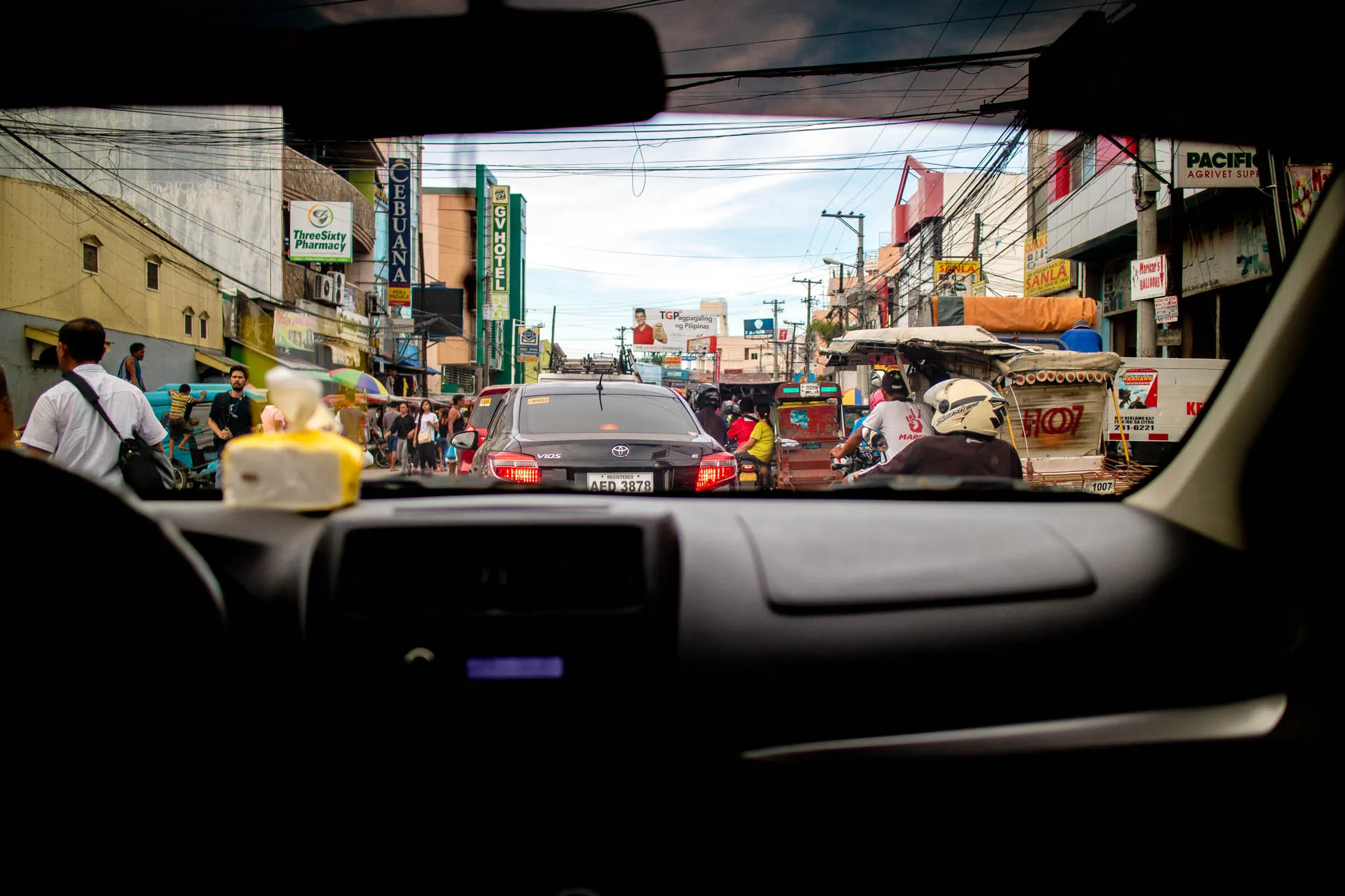 写真はフィリピンのセブで撮影されたもので、車のダッシュボード越しに街の様子を捉えています。フロントガラスの上部にはルームミラーが写り込み、そこから見える空は曇り空で、少し暗めの印象を受けます。視線の先には、電線が複雑に張り巡らされた、雑多とした街並みが広がっています。  道路は渋滞しており、乗用車やバイク、トライシクルなどがひしめき合っています。中央には黒いトヨタ・ヴィオスが写っており、ナンバープレートには「AED 3878」と記されています。周囲には人々が歩いており、商店の看板や広告などが所狭しと並んでいます。薬局「Three Sixty Pharmacy」やホテル「GV HOTEL」などの看板が見え、現地の雰囲気を伝えています。道路沿いには屋台のようなものもあり、生活感を感じさせます。全体的に活気があり、賑やかな様子がうかがえます。