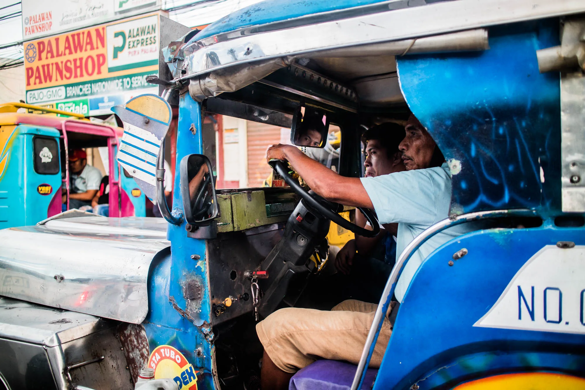 フィリピンのセブで撮影された写真です。鮮やかな青色のジプニーの運転席に、2人の男性が乗っている様子が写っています。運転手はハンドルを握り、真剣な表情で前を見据えています。助手席には若い男性が座っており、視線は同じ方向に向いています。ジプニーの内装は使い込まれた様子で、メーターパネルやステアリングホイールなどが古びています。  背景には、別のカラフルなジプニーが見えます。ピンクと水色の配色で、運転席には赤い帽子をかぶった男性が乗っています。さらに、質屋の看板が見え、通りの賑わいを伺わせます。全体的に、ローカルな雰囲気と日常生活を感じさせる一枚です。