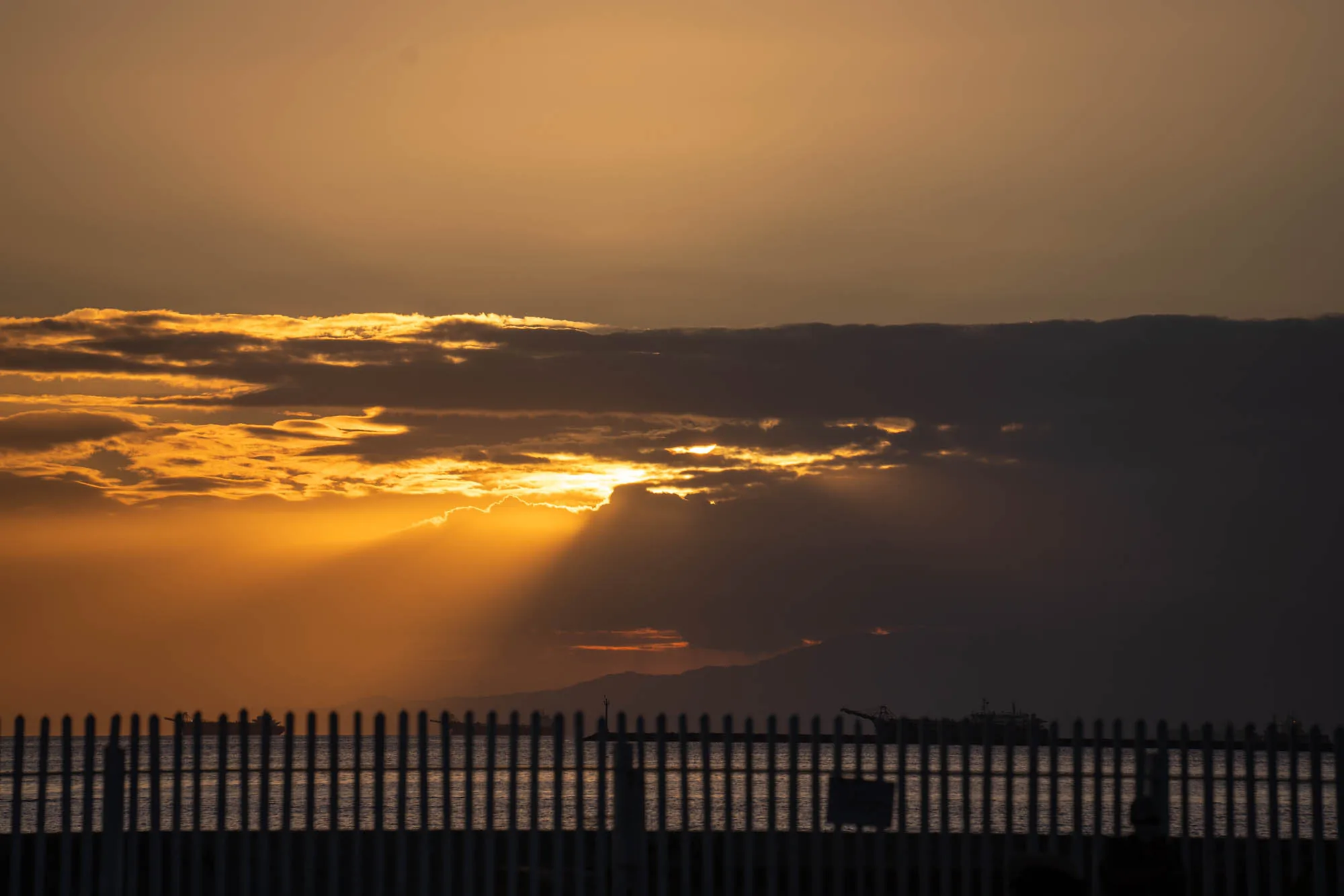 写真はフィリピンのマニラで撮影された夕焼けの風景です。手前に白い柵が並び、その向こうには海が広がっています。水平線には山並みが見え、海には大型船らしき影が確認できます。空は黄金色からオレンジ色に染まり、厚い雲の切れ間から太陽の光が力強く差し込んでいます。光芒が海面を照らし、幻想的な雰囲気を醸し出しています。全体的に落ち着いた色調で、静けさと美しさが感じられる写真です。