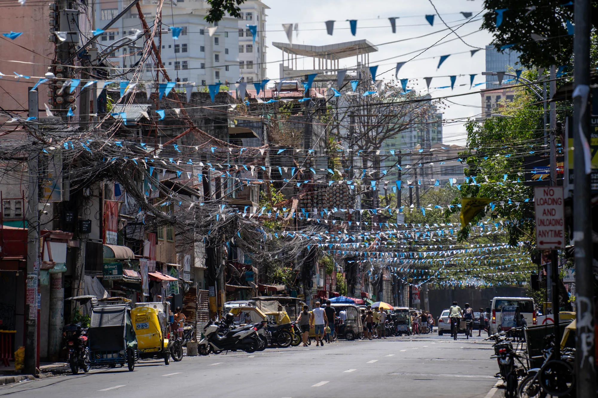 写真はフィリピンのマニラで撮影された、活気のある街並みを捉えたものです。道路の両側には建物が並び、軒先には様々な店舗が見られます。電線が複雑に絡み合い、その間には青と白の旗が飾られ、お祭りか何かのイベントの雰囲気を醸し出しています。道路にはトライシクル、バイク、車、自転車が行き交い、多くの人々が歩いています。路肩にはバイクが駐輪されており、生活感が感じられます。背景には高層ビルも見え、都市部の様子が伺えます。全体的に、マニラのエネルギーと混沌とした魅力を感じさせる一枚です。「NO LOADING AND UNLOADING ANYTIME」という標識も写っており、駐車に関するルールがあることがわかります。