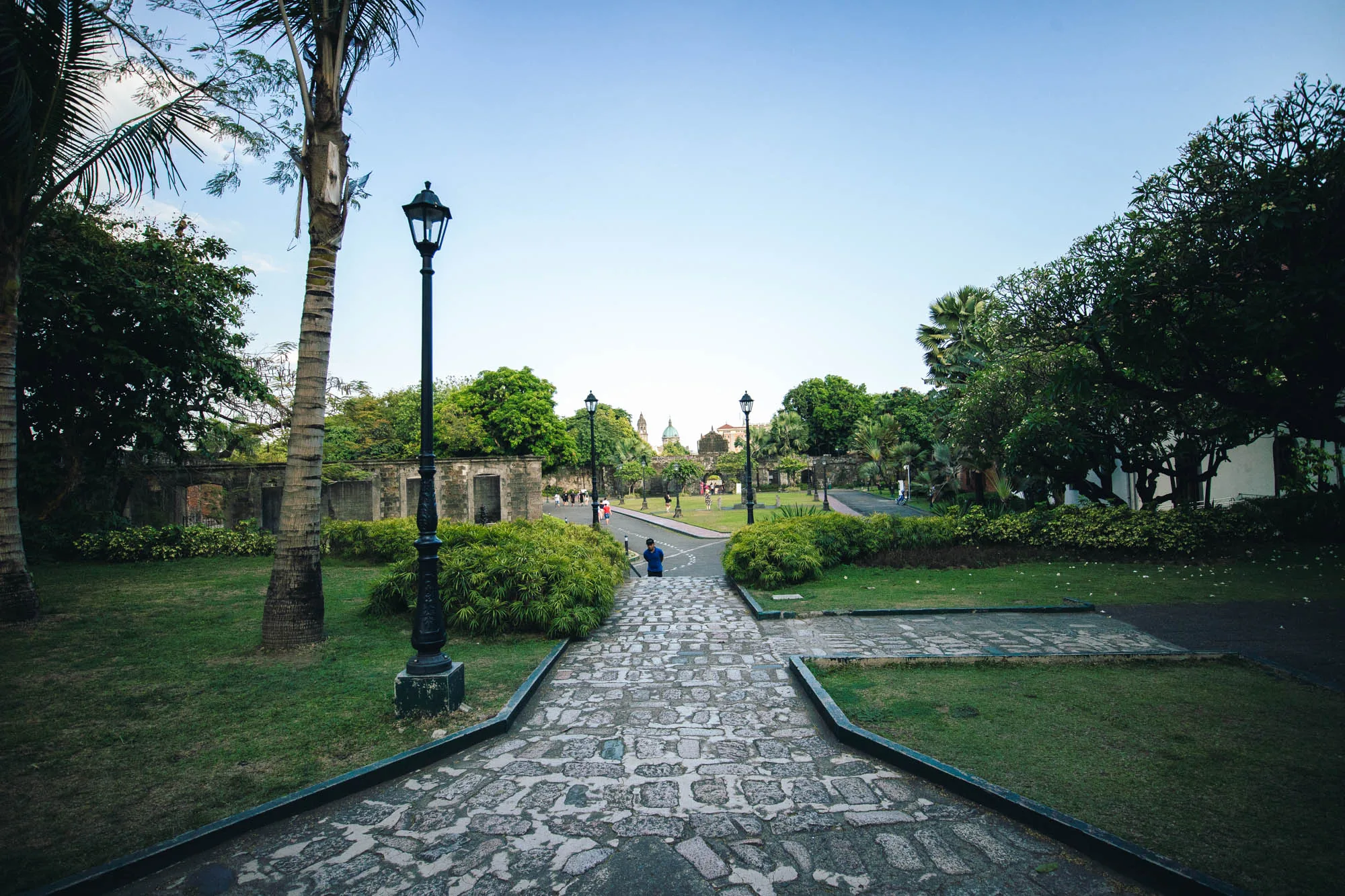 写真はフィリピンのマニラで撮影された、歴史的な公園の風景を捉えています。石畳の道が視線の先へと伸び、その先に緑豊かな公園が広がっています。道端にはレトロな街灯が立ち並び、公園内には緑の芝生や木々が生い茂り、心地よい雰囲気を醸し出しています。公園の奥には歴史的な建造物が見え、観光客らしき人々が散策している様子が伺えます。空は晴れ渡り、全体的に穏やかな時間が流れているような印象です。手前の石畳の道は少し影になっており、奥行きを感じさせる構図となっています。公園内には、ヤシの木のような熱帯の植物も見られます。