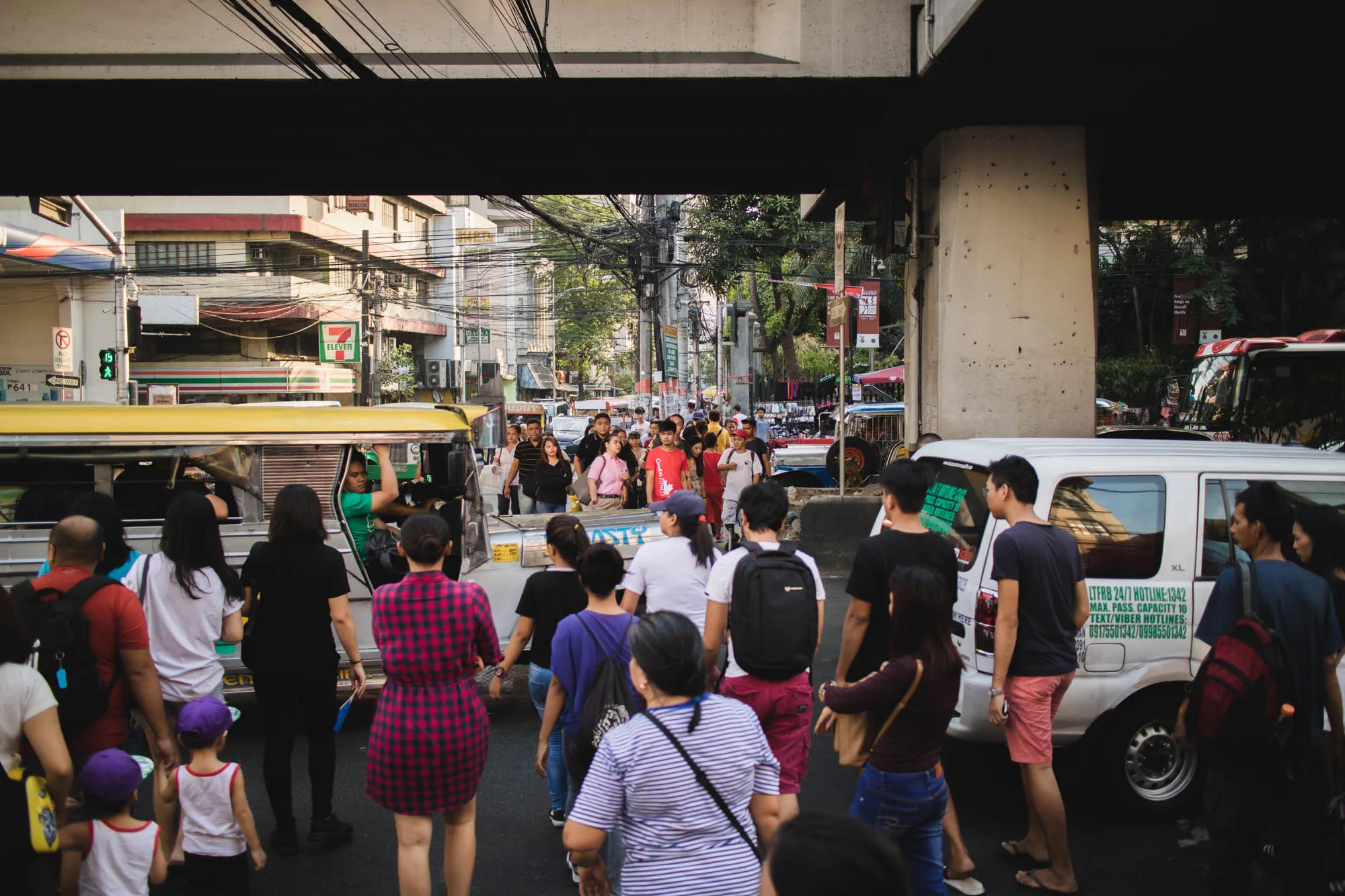 この写真はフィリピンのマニラで撮影されたもので、活気ある街の様子を捉えています。画面中央には、多数の人が道路を横断する様子が写っています。人々は様々な服装をしており、日常生活の一部を切り取ったような自然な雰囲気が感じられます。歩行者の間には、フィリピンの典型的な交通手段であるジープニーや、白いバンなどの車両も見えます。  道路の上には高架橋があり、その下を人々が通り抜けています。背景には建物が立ち並び、電線が複雑に絡み合って空を覆っています。写真全体の色調はやや暗めで、都会の喧騒と混沌とした雰囲気を伝えています。左側には「7-Eleven」の看板が見え、街の風景の一部として溶け込んでいます。全体的に、マニラの日常的な風景をリアルに表現した写真と言えるでしょう。