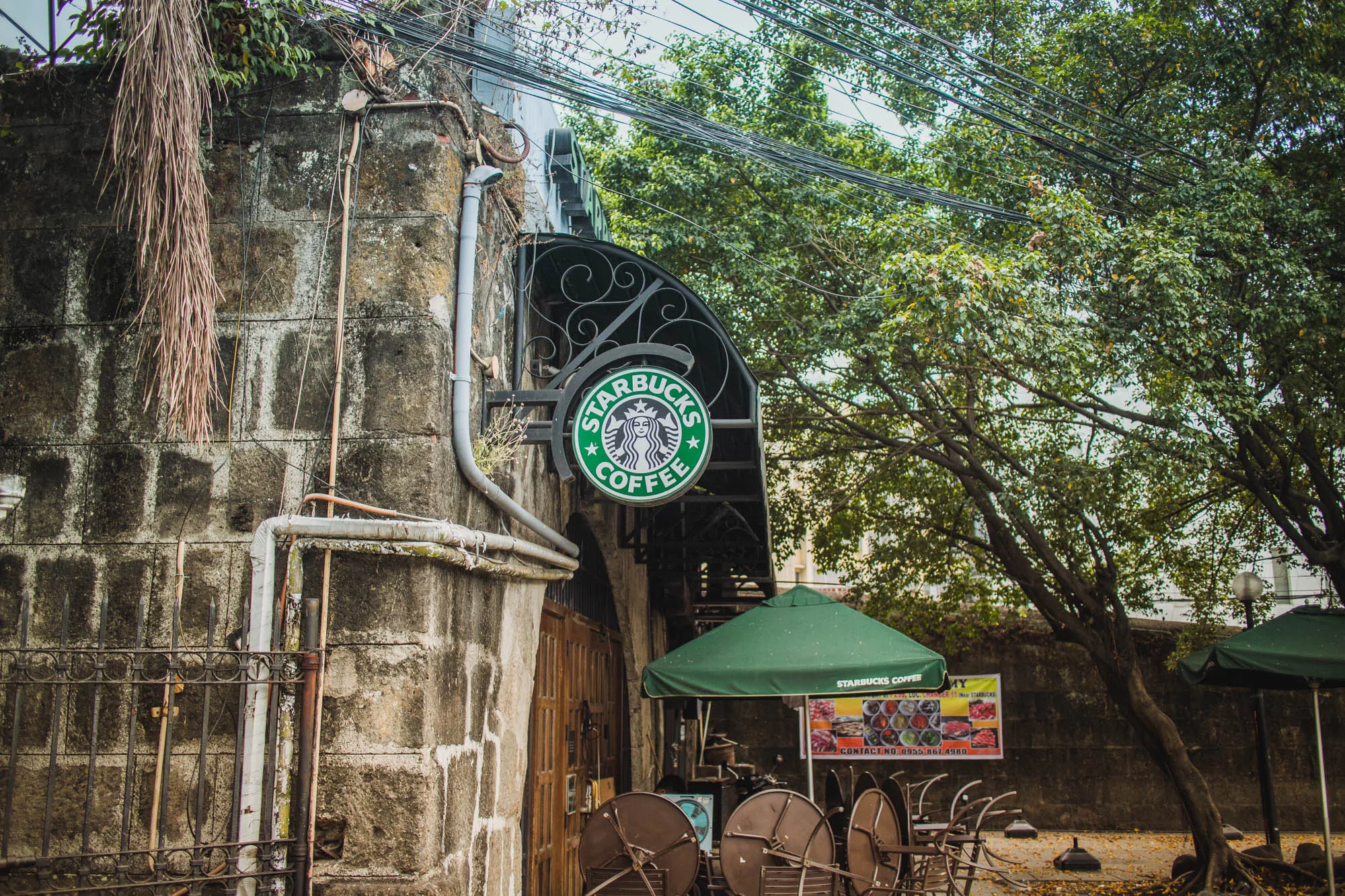 写真はフィリピンのマニラで撮影されたスターバックスの店舗の様子を捉えています。古びた石造りの壁に、装飾的な鉄製の枠で縁取られたスターバックスの緑色の丸い看板が掲げられています。壁には配管や電線がむき出しで、歴史を感じさせる風合いです。店舗の入り口には緑色のパラソルが設置され、その下にはテーブルと椅子が重ねて置かれています。パラソルの下にはメニューのポスターが掲示されています。周囲には緑豊かな木々が生い茂り、電線が空を覆っています。全体的に、歴史的な建造物と現代的なカフェが融合した独特の雰囲気を醸し出しています。
