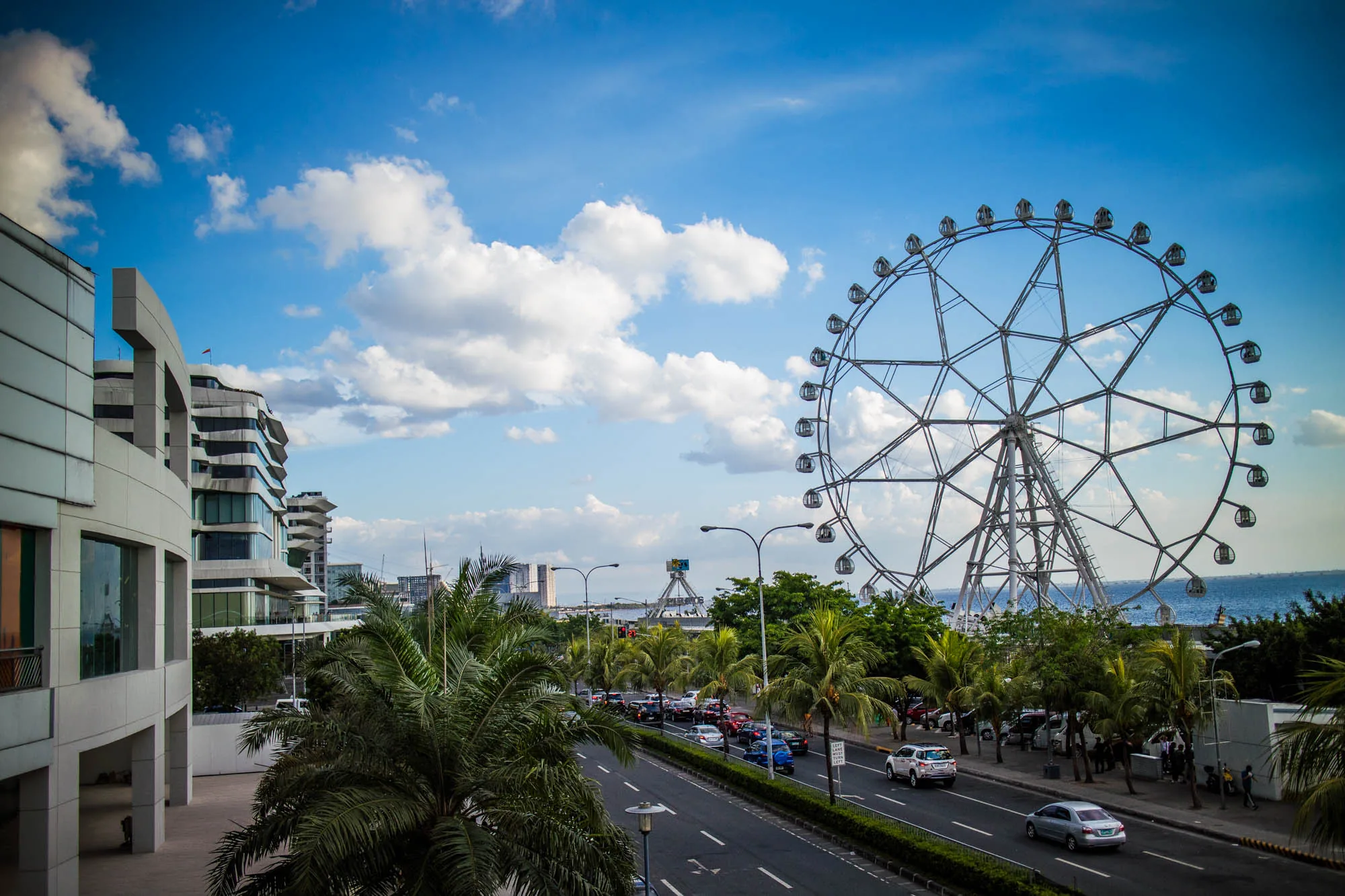 写真はフィリピンのマニラで撮影されたもので、賑やかな街並みが写っています。画面右側には大きな観覧車がそびえ立ち、その奥には海が見えます。観覧車の手前には道路があり、多くの車が行き交っています。道路沿いにはヤシの木などの緑が植えられており、熱帯の雰囲気を醸し出しています。画面左側には近代的な建物が写っており、街の発展を感じさせます。空は青く、白い雲が浮かんでおり、全体的に明るく開放的な印象を与える写真です。