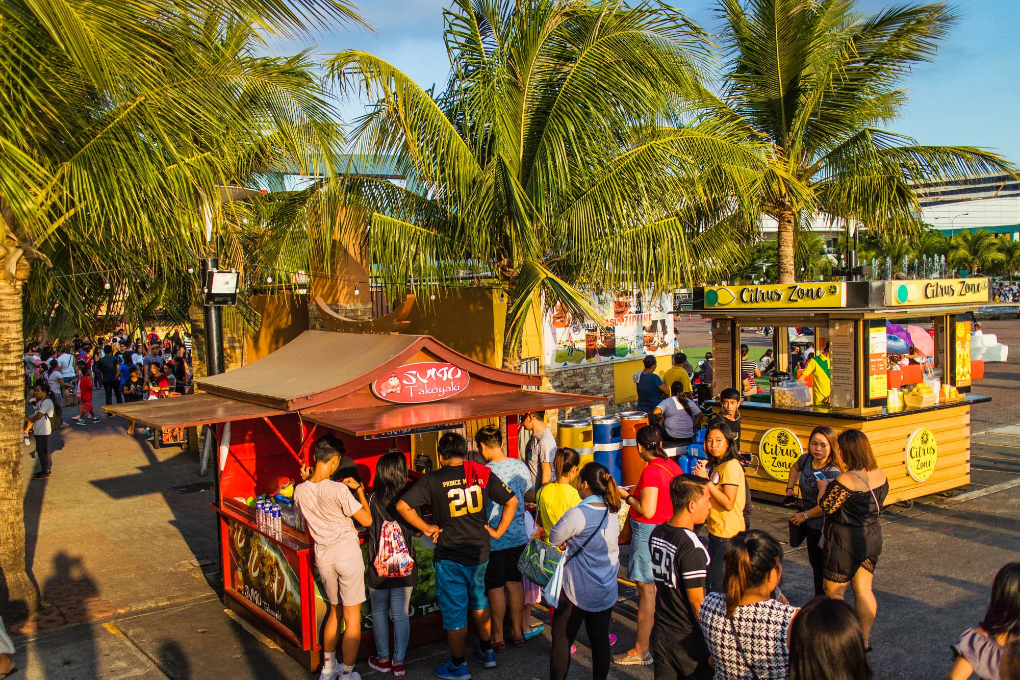 写真はフィリピンのマニラで撮影されたもので、屋台が立ち並び、多くの人々で賑わっている様子が写っています。手前には赤い屋根の「SUGO Takoyaki」と書かれたたこ焼き屋の屋台があり、多くの人が列を作って並んでいます。その隣には黄色い「Citrus Zone」と書かれた柑橘系の飲み物などを販売していると思われる屋台があります。その周囲にも人が集まっています。  背景には多くのヤシの木がそびえ立ち、トロピカルな雰囲気を醸し出しています。また、遠くには近代的な建物が見え、マニラの都市部であることがわかります。多くの人々が様々な服装をしており、活気と多様性のある様子が伝わってきます。全体的に明るく暖かい光が差し込み、楽しげな雰囲気を強調しています。昼間の屋外で、多くの人々が食べ物を求めて集まる様子が写っており、賑やかな観光地の風景を捉えた一枚です。
