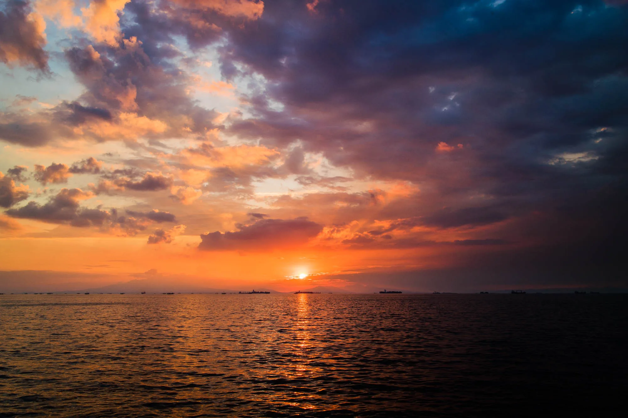 写真はフィリピンのマニラで撮影された夕焼けの海の風景です。空は、オレンジ、ピンク、紫、そして濃い青色の雲で彩られており、燃えるような夕日が地平線に沈む瞬間を捉えています。太陽の光が海面に反射し、キラキラと輝く光の道を作り出しています。水平線には、いくつかの船が見え、遠くの島影のようなものも確認できます。全体的に、ドラマチックで美しい自然の光景が表現されています。