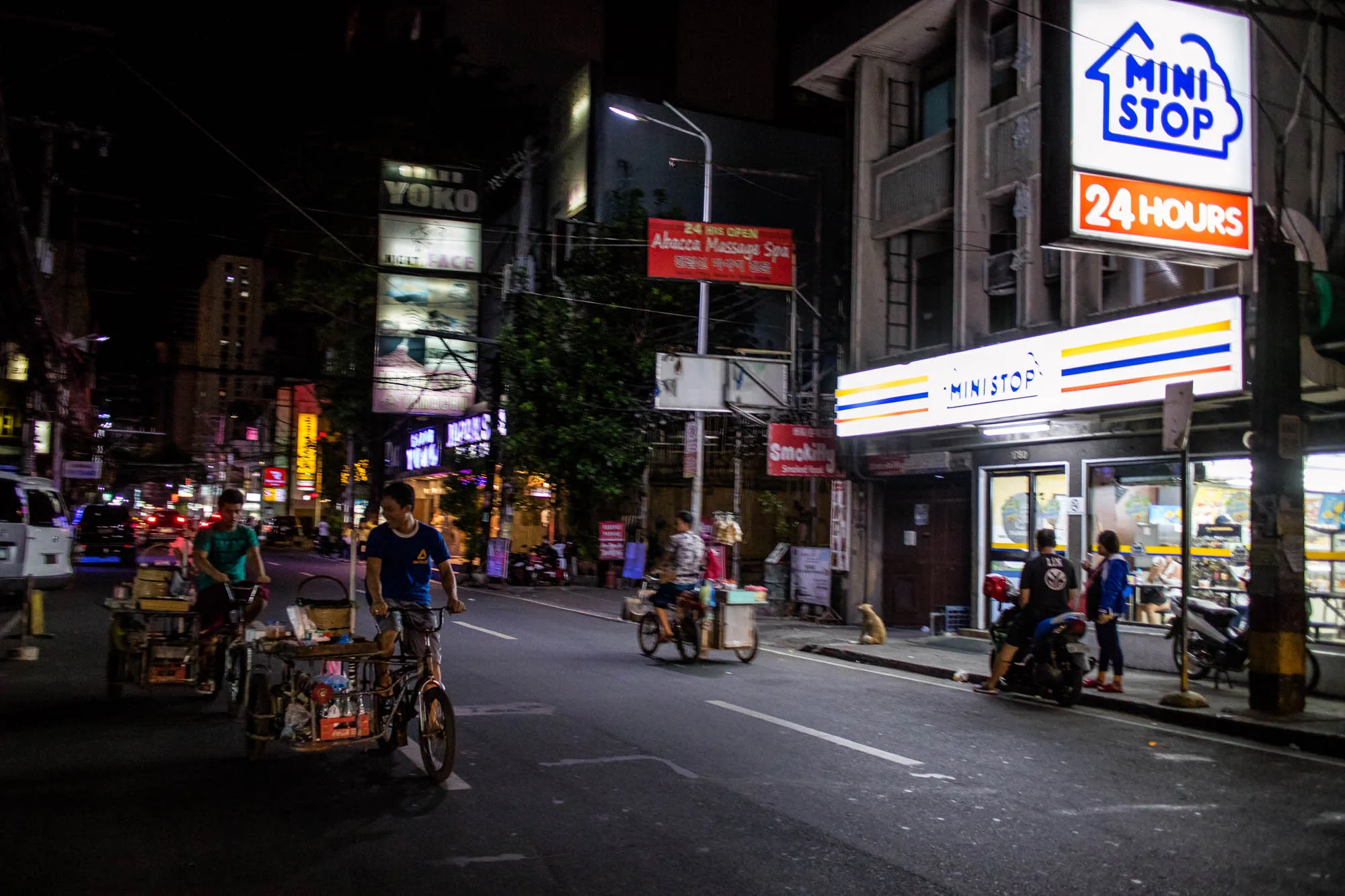 フィリピンのマニラで撮影された夜の写真です。画面右側には、明るく照らされた「MINISTOP」の看板が見えます。その下の店舗の前には、バイクが数台停まっており、人々がたむろしています。道路には、手作りの屋台を引いた自転車に乗る人々が数人写っています。背景には、「YOKO」などのネオンサインや、その他の店舗の明かりがぼんやりと見え、賑やかな夜の街の雰囲気を醸し出しています。道路脇には野良犬のような犬が座っています。全体的に、活気がありながらも少し薄暗い、東南アジアの都市の夜の風景を切り取った写真です。
