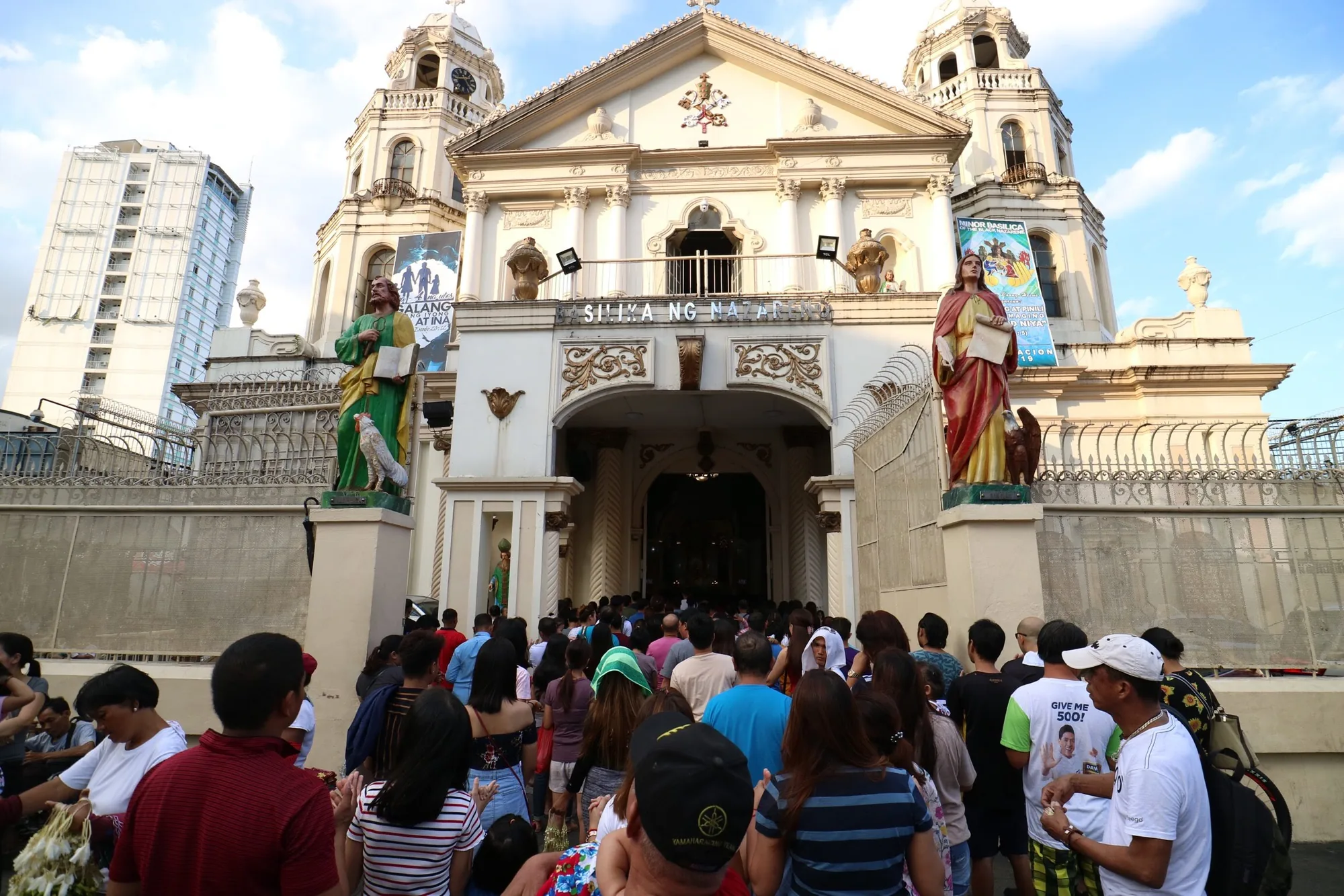 フィリピンのマニラにある教会（おそらくミノール・バシリカ）の前で撮影された写真です。教会の入り口には多くの人が群がっており、中に入ろうとしている様子がうかがえます。 教会の建物は白く、精巧な装飾が施されています。入り口の両側には聖人の像が立っており、建物の壁にはイベントのバナーが掲げられています。背後には近代的な高層ビルが見えます。群衆の中にはさまざまな服装をした人がおり、一部の人は何かを売っているように見えます。全体的に、宗教的な行事やイベントのために人々が集まっている賑やかな様子が伝わってきます。