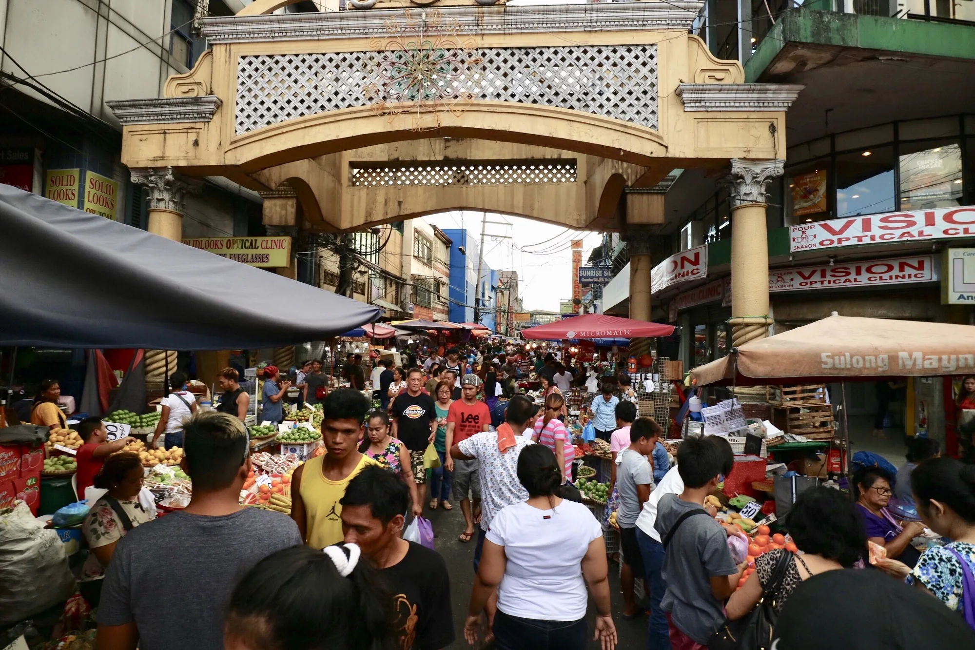 写真はフィリピンのマニラで撮影された、活気ある市場の様子を捉えています。アーチ状の装飾が施された入り口をくぐると、多くの人々で賑わう通りが見えます。通りには、野菜や果物などを売る露店が所狭しと並び、買い物客で混雑しています。露店のテントや傘が日差しを遮り、通りはやや暗い印象です。人々の服装は様々で、Tシャツやカジュアルなシャツを着ている人が多いようです。写真全体から、現地の生活感が伝わってくるような、活気のある雰囲気を感じられます。看板には「Optical Clinic」といった文字が見られ、お店が混在している様子が伺えます。