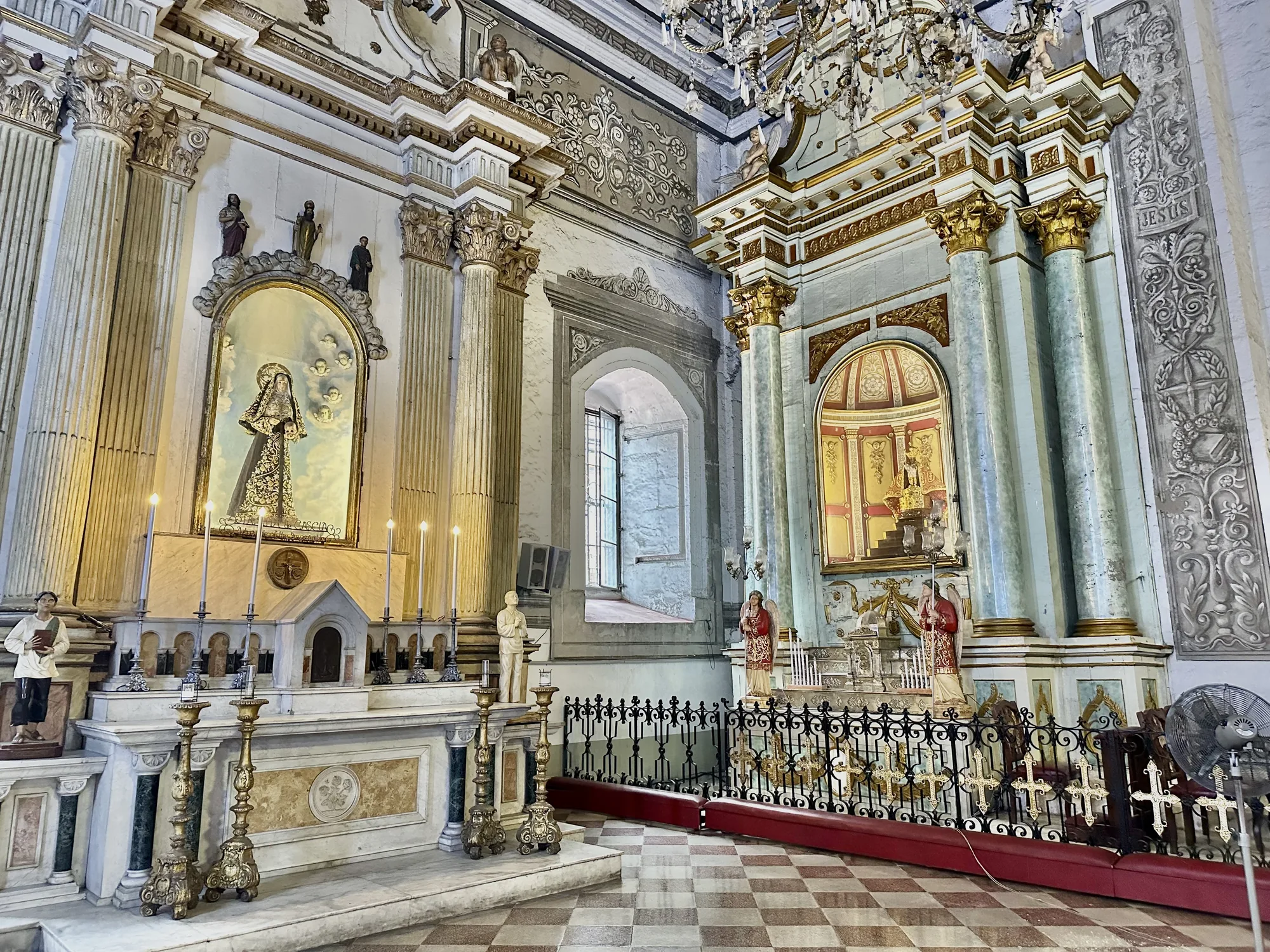 写真はフィリピン、マニラの教会内部を写したものです。非常に装飾的で、歴史を感じさせる雰囲気があります。床は茶色と白のチェック柄で、空間全体は石造りのように見えます。  左側には祭壇があり、聖母像が飾られています。聖母像の周りには小さな天使の絵が浮かんでおり、祭壇には蝋燭が灯されています。祭壇の脇には人物の像が置かれています。  右側にも同様に装飾された祭壇が見えます。こちらも天使像が両脇に配置され、祭壇の前には黒い鉄柵が設けられています。柵には金色の十字架が飾られています。  壁には精緻な模様が描かれており、柱は白と金色で装飾されています。全体的に豪華で荘厳な雰囲気であり、歴史的な価値を感じさせます。