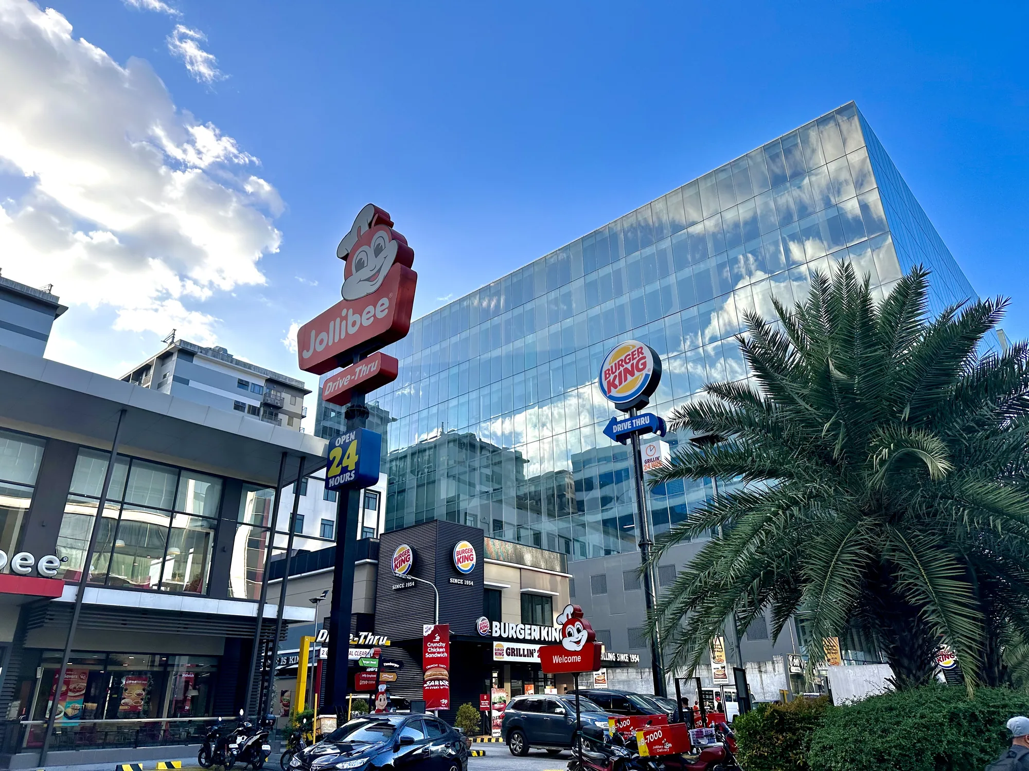 写真はフィリピンのマニラで撮影されたもので、青空の下、賑やかな街の様子が写っています。前景には、フィリピンの代表的なファストフードチェーン「ジョリビー（Jollibee）」と「バーガーキング（Burger King）」の店舗が並んでいます。ジョリビーの赤いキャラクターが特徴的な看板や、24時間営業を示す表示が目を引きます。バーガーキングの看板も同様に目立っています。店舗の前には車やバイクが停まっており、活気のある様子が伝わってきます。背景には、ガラス張りの近代的な高層ビルがそびえ立ち、空の雲が反射して美しい景観を作り出しています。ヤシの木が南国らしい雰囲気を添え、マニラの都市景観を象徴する一枚と言えるでしょう。