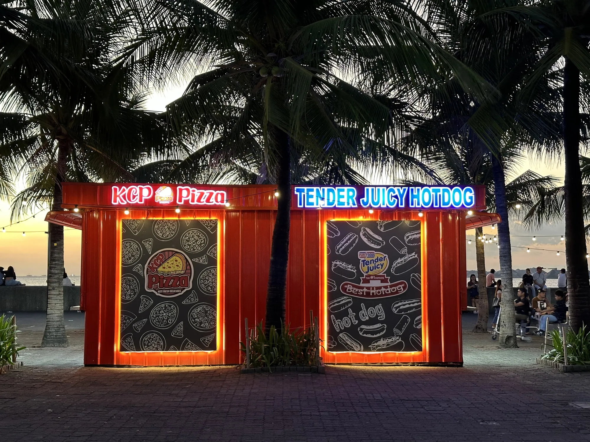 写真はフィリピンのマニラで撮影されたもので、夕暮れ時の風景が写っています。手前にはオレンジ色のコンテナで作られた屋台が2つ並んでおり、左側には「KCP Pizza」、右側には「TENDER JUICY HOTDOG」とネオンサインで書かれています。屋台の側面にはピザとホットドッグのイラストが描かれた黒板のような看板が飾られています。  屋台の後ろにはヤシの木が並び、夕日の光を通してシルエットになっています。さらに奥には海が見え、その手前の遊歩道には人々が座ってくつろいでいる様子が写っています。全体的に、夕暮れ時の穏やかな雰囲気と、屋台のネオンサインの明るさが印象的な写真です。