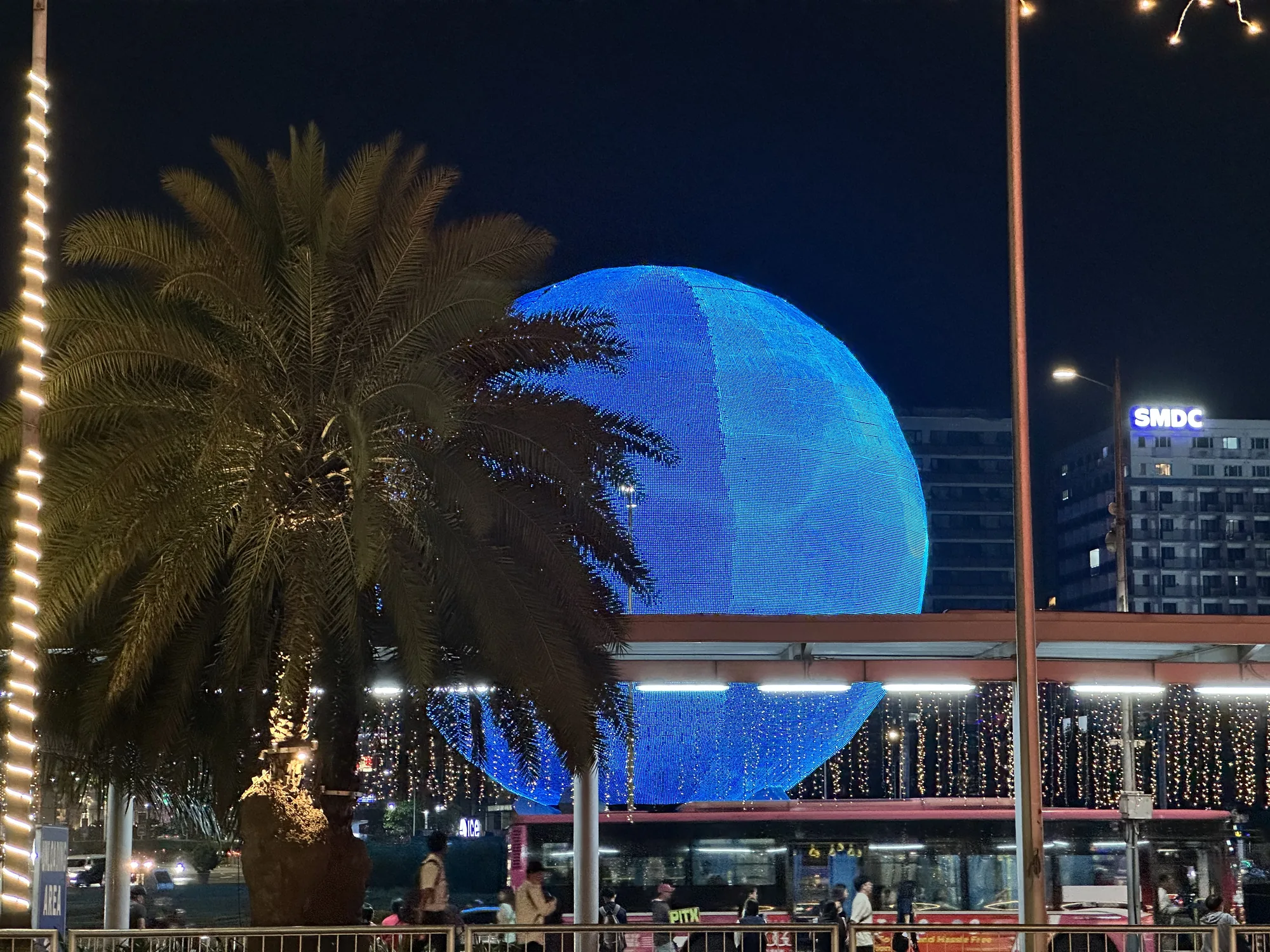 写真はフィリピンのマニラで撮影された夜景です。画面中央には、青色のイルミネーションで飾られた巨大な球体が目を引きます。球体の表面は無数の小さな光で覆われており、夜空に浮かび上がるような幻想的な雰囲気を醸し出しています。球体の手前にはヤシの木があり、幹には黄色のイルミネーションが巻き付けられています。ヤシの葉が球体を部分的に覆い、奥行きのある構図になっています。  写真の下部にはバス停のような場所があり、数人の人々がバスを待っている様子が伺えます。バスの車体には広告が描かれており、街の喧騒を感じさせます。背景には高層ビル群がそびえ立ち、そのうちの1つの建物には「SMDC」の文字が光っています。街灯や建物の光が夜空を照らし、全体的に暖かく賑やかな雰囲気が漂っています。