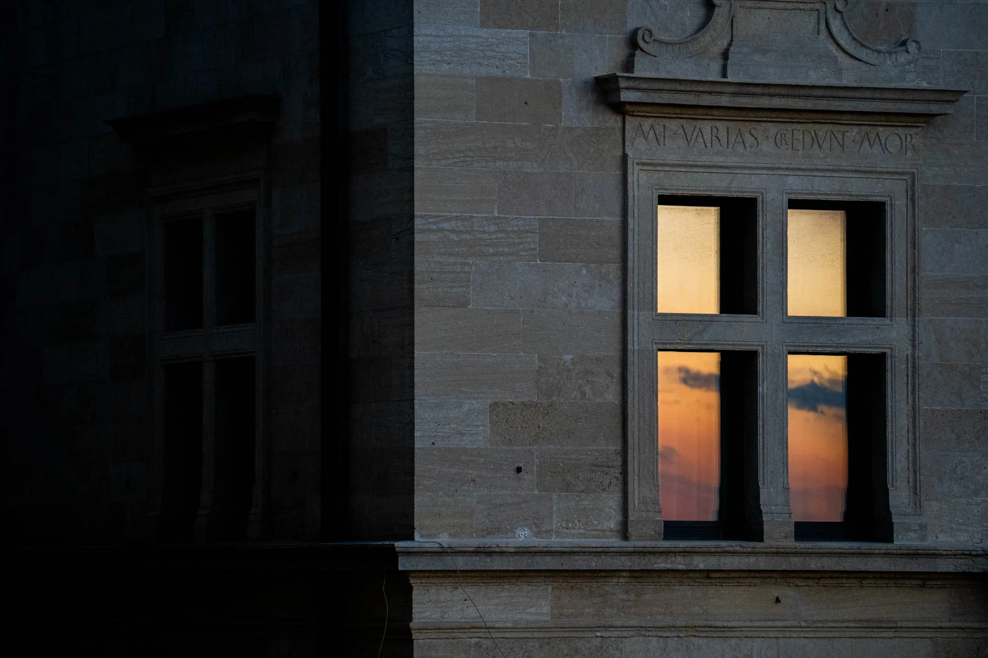 サンマリノで撮影されたと思われるこの写真は、石造りの建物の壁面と窓を捉えています。画面左側は深い影に覆われており、窓の一部がかすかに見える程度です。一方、画面右側は比較的明るく、窓枠の装飾や石壁の質感がはっきりと見て取れます。特に目を引くのは窓ガラスに映る夕焼けの光景です。オレンジ色と青色のグラデーションが美しく、空に浮かぶ雲の形も認識できます。窓枠の上部には文字が刻まれており、建物の歴史を感じさせます。全体的に落ち着いた色調で、静かで荘厳な雰囲気が漂っています。夕暮れ時のノスタルジーや、歴史的な建造物の重厚感を表現した写真と言えるでしょう。