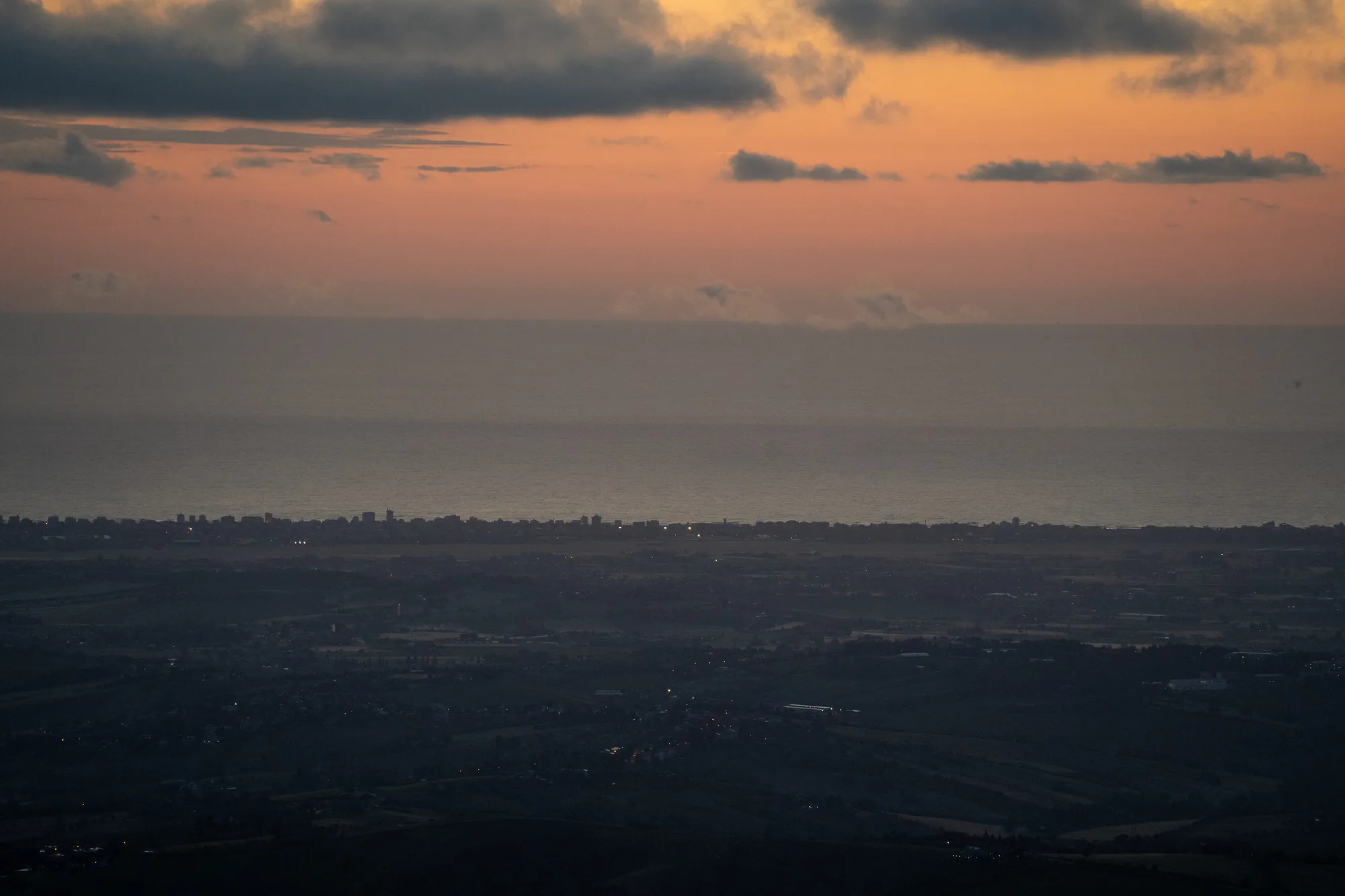写真は、サンマリノで撮影された夕暮れ時の風景です。空はオレンジ色からピンク色にグラデーションを描き、上空には濃い灰色の雲が点在しています。水平線には、ぼんやりとした海の景色が広がっており、海岸線に沿って建物が並んでいるのが見えます。海岸線の手前には、暗い緑色の丘陵地帯が広がっており、小さな集落の明かりが点々と見られます。全体的に、穏やかで静かな雰囲気が感じられます。夕暮れ時の光と影のコントラストが、風景に深みを与えています。