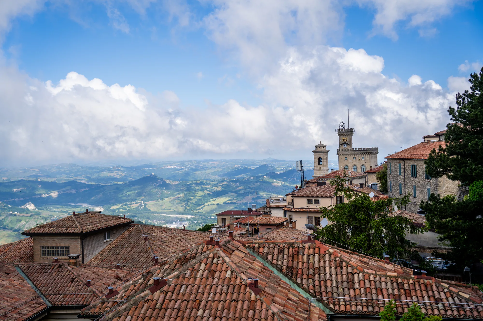 写真は、サンマリノの古い街並みを上から見下ろすように撮影されたものです。手前にはオレンジ色の瓦屋根が連なり、歴史を感じさせる風景が広がっています。屋根の奥には、ベージュ色の建物や石造りの建物、そして特徴的な時計台が見えます。  背景には、緑豊かな丘陵地帯が広がり、遠くまで見渡せる絶景です。空には白い雲が浮かび、青空とのコントラストが美しいです。全体的に明るく開放的な雰囲気で、サンマリノの美しい自然と歴史的な街並みが調和している様子が伝わってきます。