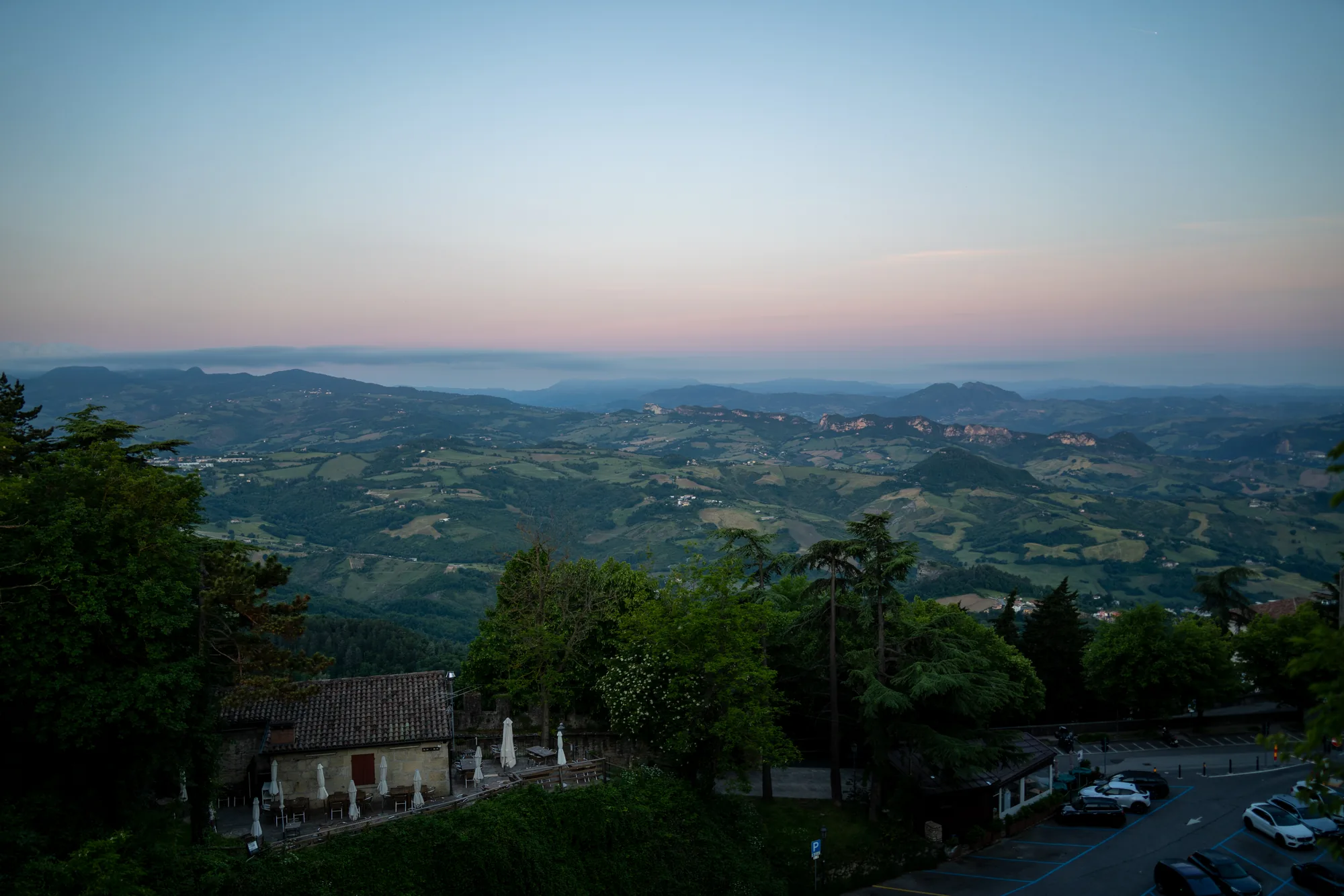 写真はサンマリノの不明な場所で撮影された、夕暮れ時の風景を捉えています。前景には、レンガ屋根の建物と、そのテラスに置かれたテーブルと椅子が並ぶレストランが見えます。建物とテラスは緑豊かな木々に囲まれており、その奥には駐車場があり、数台の車が駐車されています。  写真の中央部分には、起伏に富んだ丘陵地帯が広がっており、緑の畑や木々がパッチワークのように景色を彩っています。遠景には、霞がかった山々が連なり、空は上部が薄い青色から、地平線に向かってピンク色へとグラデーションを描いています。夕暮れ時の柔らかい光が、風景全体を穏やかで幻想的な雰囲気に包んでいます。