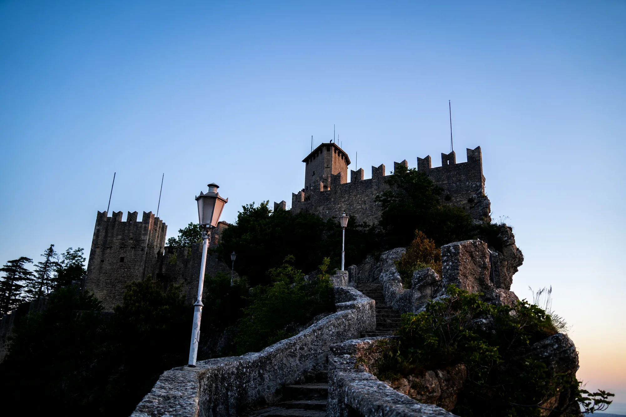 写真には、サンマリノにある古城が写っています。城は岩山の上に建てられており、石造りの壁や塔が特徴的です。空は澄み切った青色で、夕暮れ時なのか、地平線近くはオレンジ色に染まっています。城へと続く石段があり、両脇には石造りの手すりがあります。石段のそばには街灯が立っており、古城の雰囲気を引き立てています。城の周囲には木々が生い茂っており、自然豊かな環境であることが伺えます。全体的に静かで落ち着いた雰囲気の写真です。