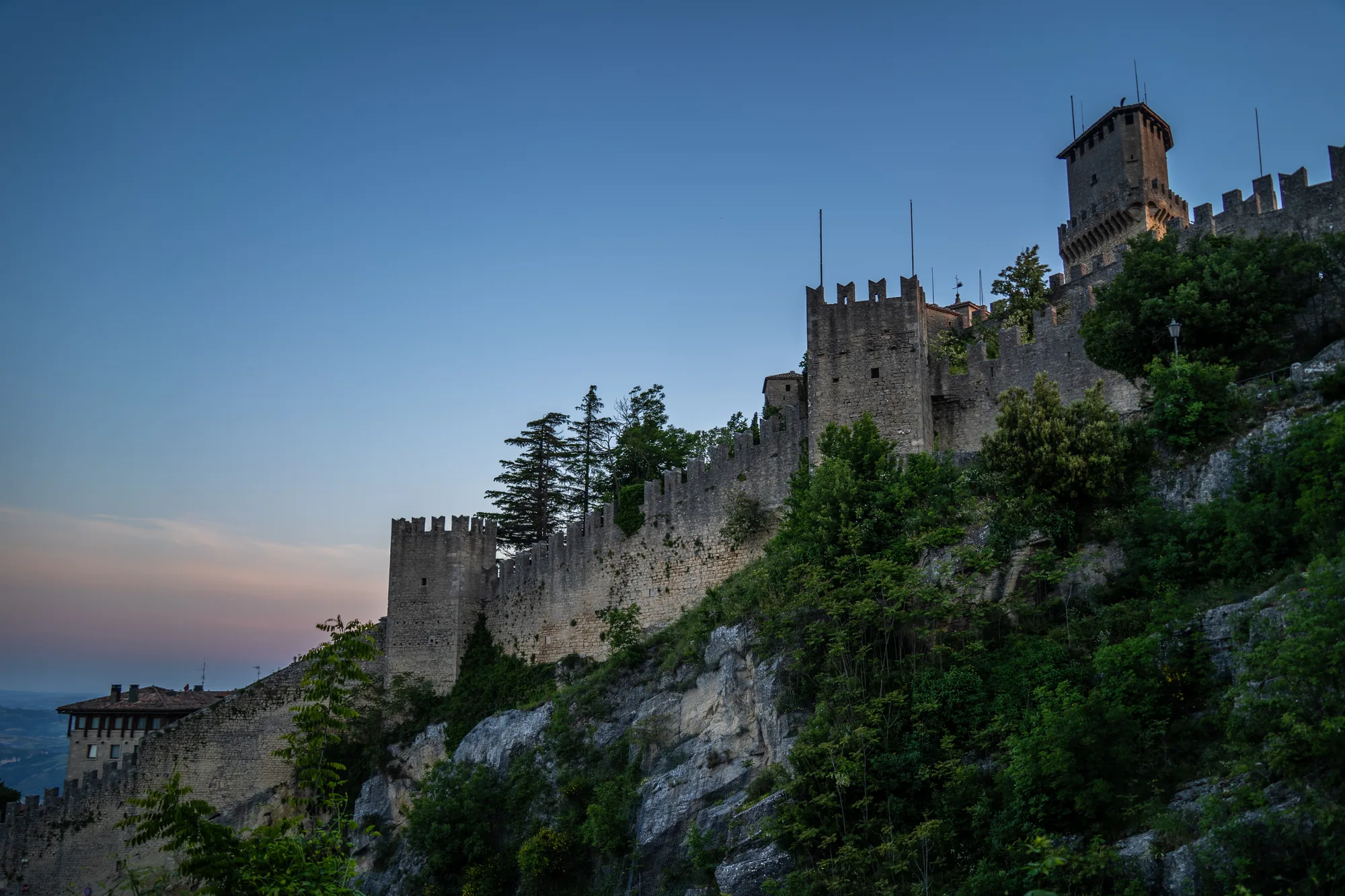 写真には、サンマリノにある要塞都市が写っています。石造りの壁と塔が崖の上にそびえ立ち、歴史を感じさせる風景です。空は夕暮れ時のグラデーションで、オレンジ色から青色へと変化しています。要塞の周りには緑豊かな木々が生い茂り、自然と建造物が調和している様子が伺えます。写真全体的に、静かで荘厳な雰囲気が漂っています。