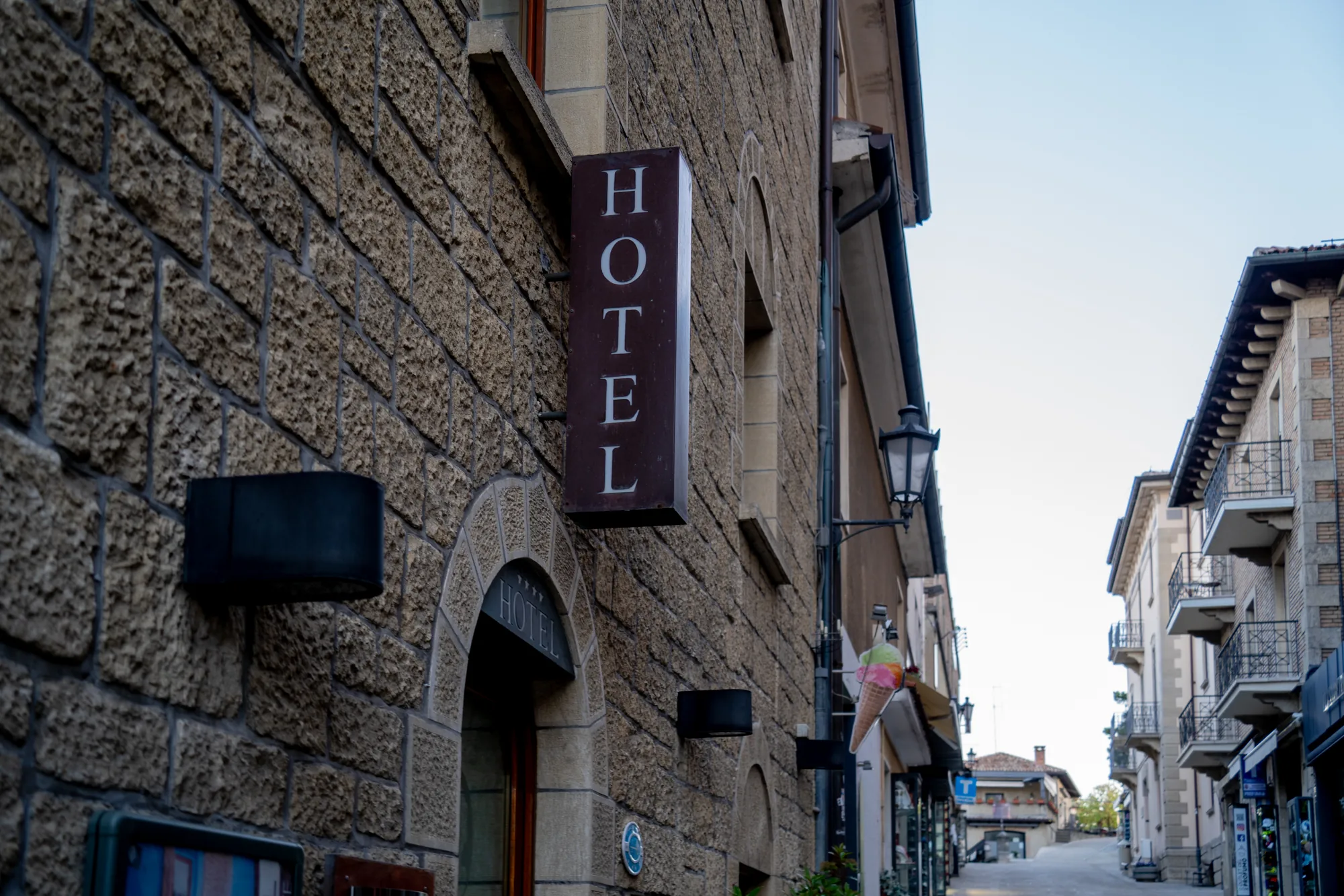 写真は、サンマリノの細い通りに面したホテルの外観を捉えています。建物は石造りで、重厚感のある壁が特徴的です。入口の上にはアーチ状の装飾があり、「HOTEL」と刻まれた看板が掲げられています。その上には縦長の「HOTEL」と書かれた看板が掲げられています。通りの奥には、バルコニー付きの建物が並び、歴史的な街並みを形成しています。空は明るく、晴天の日の撮影であることがわかります。通りには人通りは少ないようで、静かで落ち着いた雰囲気が漂っています。ホテルの外壁には照明器具が取り付けられており、夜には暖かく照らされることが想像できます。全体的に、サンマリノの美しい景観とホテルのクラシカルな雰囲気が調和した写真です。