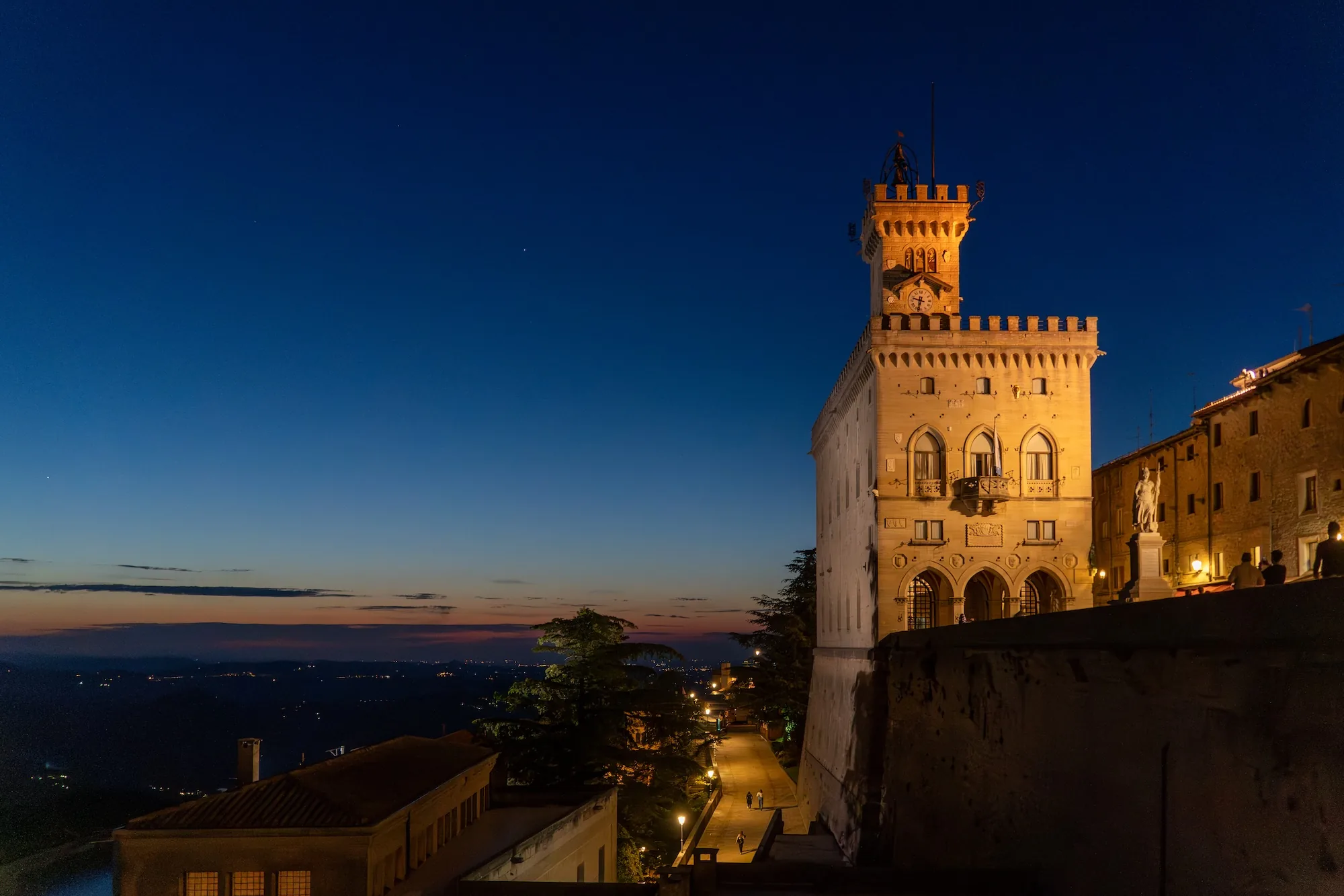 写真はサンマリノで撮影された夜景です。空は深い青色からオレンジ色のグラデーションに染まり、遠くの地平線には街の灯りが輝いています。手前には、ライトアップされた歴史的な建造物、特に時計塔が印象的です。塔は石造りで、細部まで丁寧に装飾されています。塔の横には他の建物が並び、道には街灯が灯り、人々が歩いている様子が見えます。写真全体は静かで穏やかな雰囲気を醸し出しています。