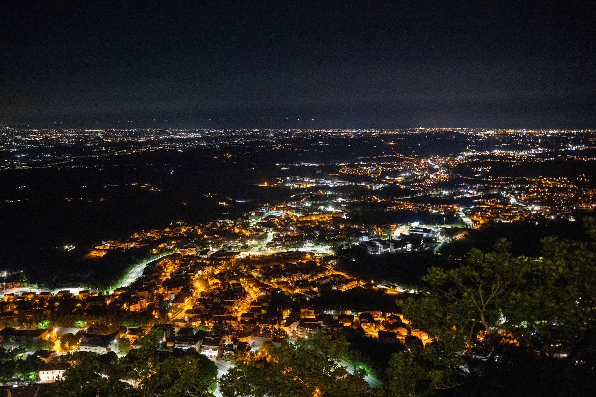 写真はサンマリノで撮影された夜景です。暗い空の下、広大な都市がオレンジ色の光で輝いています。手前には木々の緑が配置されており、遠近感を強調しています。都市の灯りは、まるで無数の星が地上に降りてきたかのように、暖かくきらびやかです。写真全体は静かで落ち着いた雰囲気で、夜の都市の美しさを捉えています。