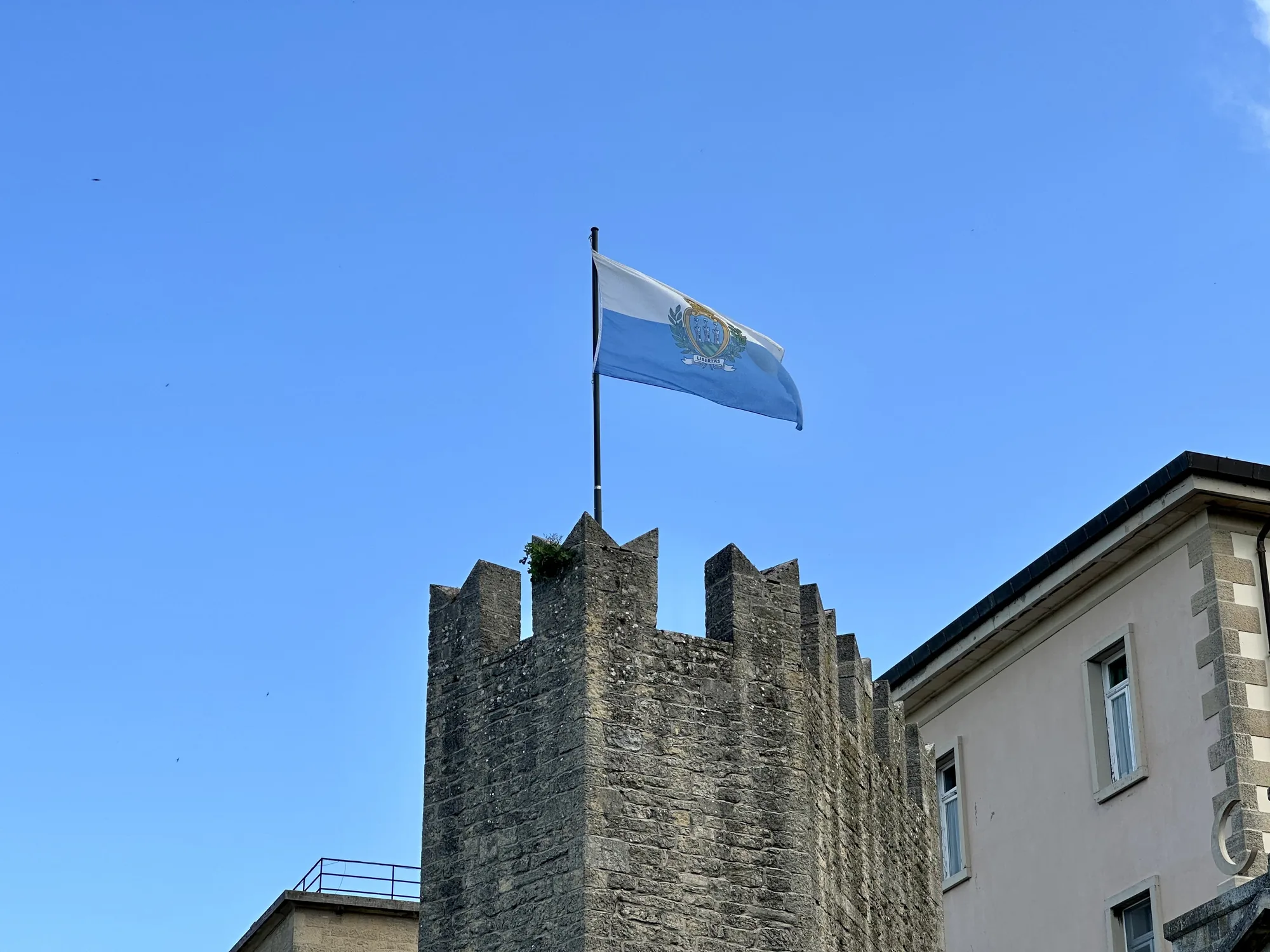 青空を背景に、サンマリノの国旗が石造りの塔の上に掲げられている写真です。旗は白と水色の二色旗で、中央には国章が描かれています。塔は石積みでできており、上部にはギザギザの稜線があります。塔の頂上付近には、わずかに草が生えているのが見えます。塔の隣には、ベージュ色の建物の一部が見えており、窓がいくつか配置されています。空は澄み渡っており、平和で穏やかな雰囲気を醸し出しています。全体的に、サンマリノの象徴的な風景を捉えた写真です。