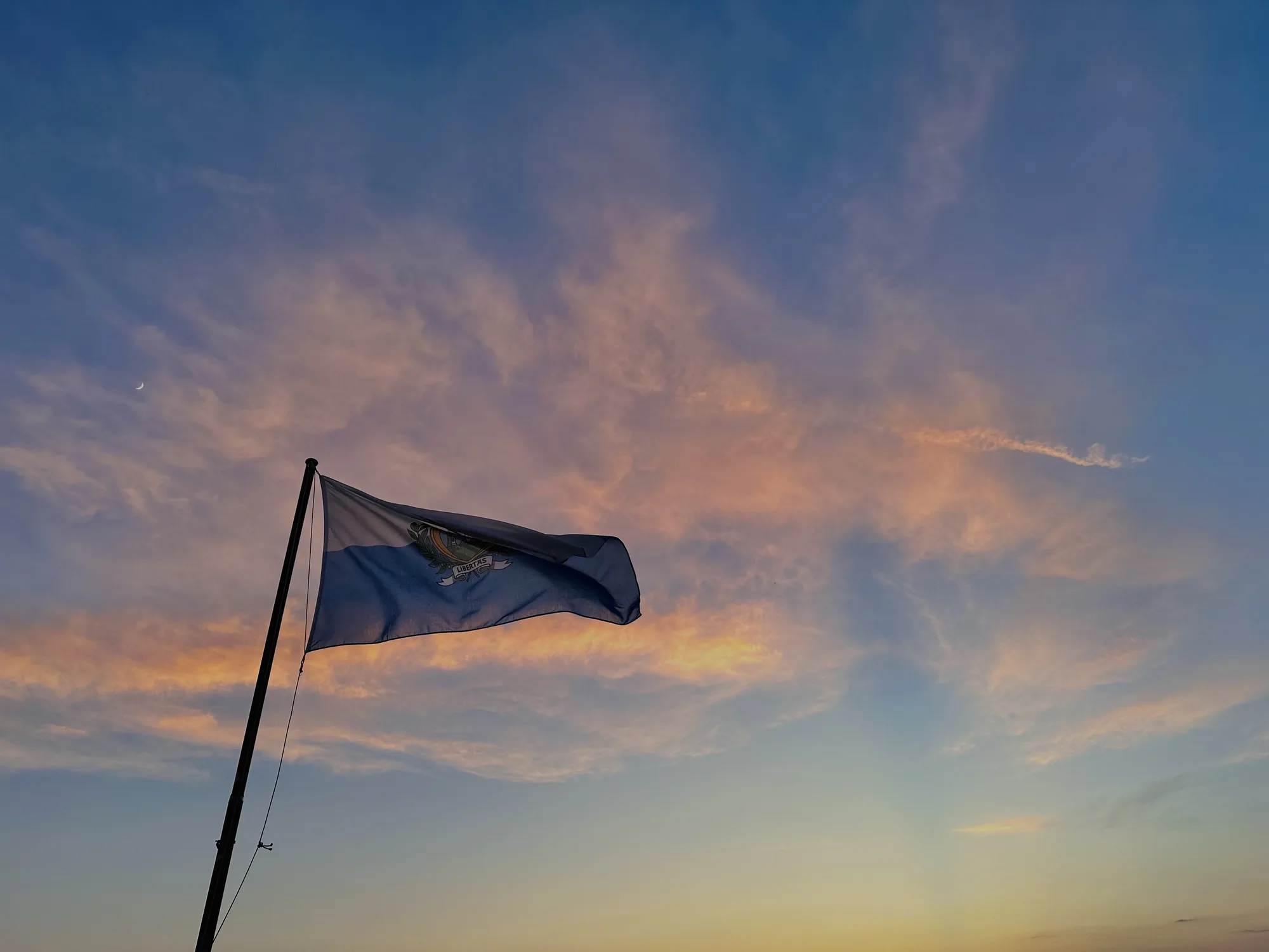 写真には、サンマリノの旗が夕焼け空を背景に掲げられている様子が写っています。旗は青と白のツートンカラーで、中央には国の紋章が描かれています。空は、夕日の光を浴びてピンクやオレンジ色に染まり、薄い雲がたなびいています。画面の左上には、三日月が小さく見えています。空の色は上部では濃い青色で、下部に向かって徐々に明るい黄色へと変化しています。旗のポールは黒く細長く、旗を支えています。全体的に、静かで平和な雰囲気の写真です。