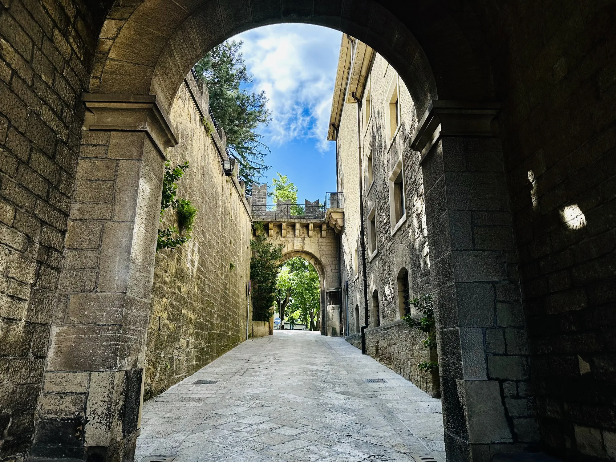 写真はサンマリノで撮影されたと思われる、歴史を感じさせる石造りの街並みを捉えたものです。写真の中心にはアーチ状の通路があり、その先にも別のアーチが見え、奥へと道が続いています。両側の建物も同じく石造りで、古い石畳の道との組み合わせが、重厚感と趣のある雰囲気を醸し出しています。空は晴れており、白い雲が浮かんでいます。アーチの影になっている部分と、日が当たっている部分のコントラストが美しく、歴史的な建造物の質感を際立たせています。全体的に静かで落ち着いた印象を受け、観光客が少ない時間帯に撮影されたかのような雰囲気が感じられます。