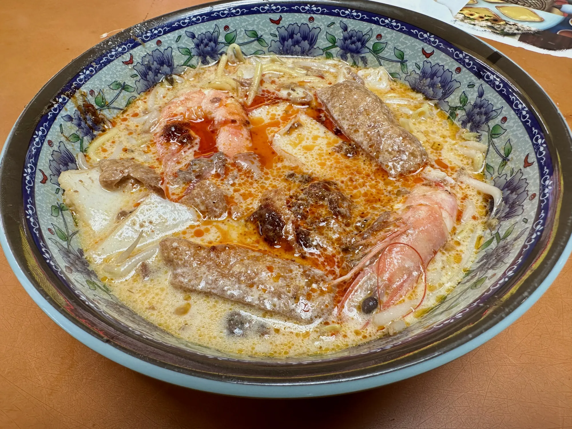 シンガポールで撮影されたと思われるラクサの写真です。ラクサは、ココナッツミルクベースのスープに麺、エビ、揚げ豆腐、魚介類、香辛料などを加えた料理です。写真に写っているラクサは、大きめの青い模様の入った器に盛られており、濃厚なココナッツミルクのスープに、赤く染まったラー油のようなものが浮かんでいます。具材として、殻付きのエビが2尾、揚げ豆腐と思われるものが数個、魚介類や肉団子のようなものも見えます。麺はスープに隠れて見えにくいですが、おそらく太めの麺が使用されているでしょう。全体的に濃厚で食欲をそそる見た目です。テーブルの質感から、ローカルなレストランで撮影された可能性が高いと思われます。